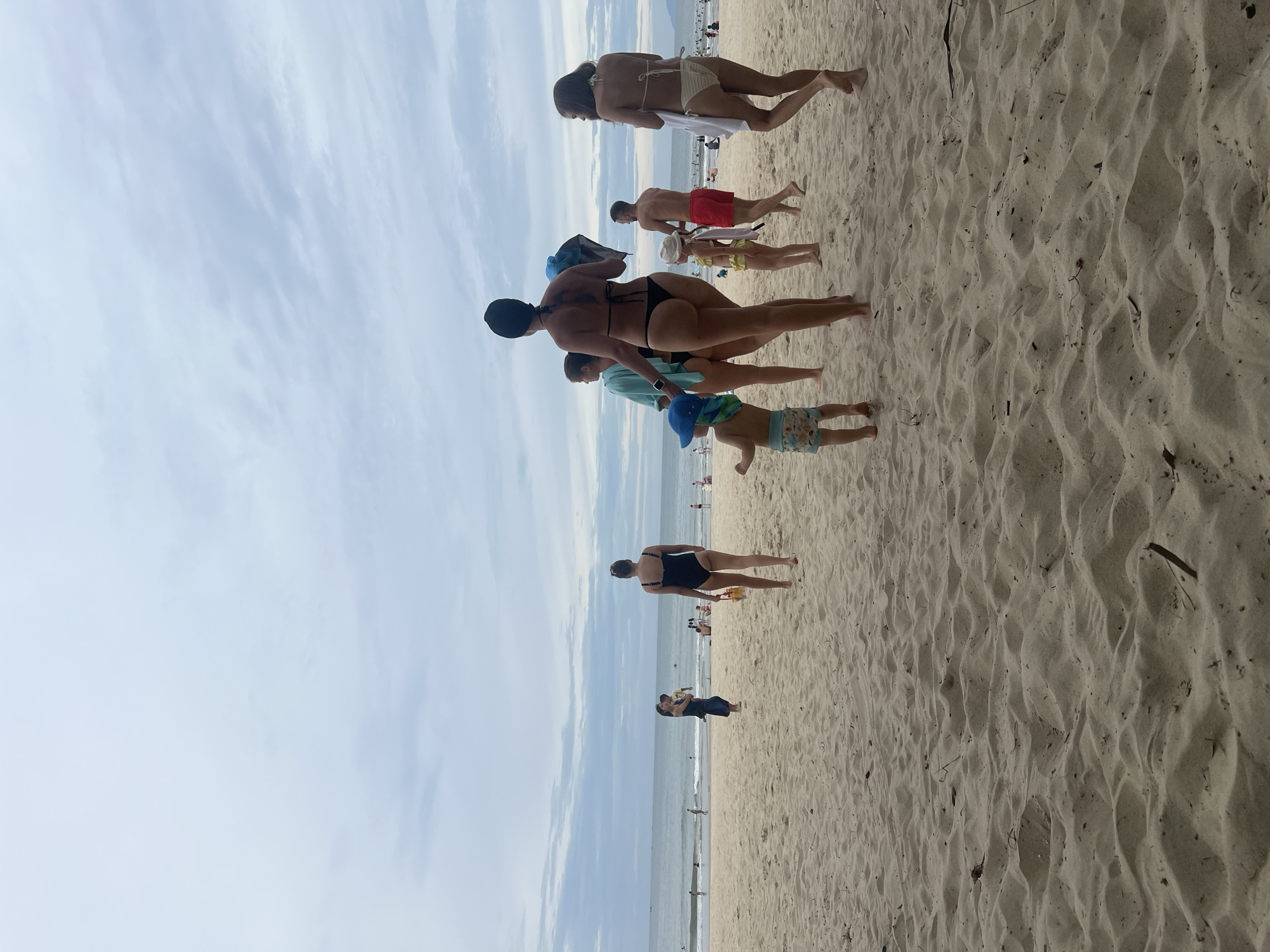 The image shows a crowded beach scene in Da Nang, Vietnam, with the ocean in the background and a sandy foreground. Several groups of people are visible, all wearing swimwear. In the foreground, a woman in a black bikini walks away from the camera, carrying a yellow bucket and a blue towel. To her left, a child in a blue hat and patterned swim trunks walks towards the left. Behind them, a woman in a blue cover-up walks away from the camera, and a woman in a black swimsuit is also walking away. Further to the left, a woman in a dark blue dress holds a child. In the middle ground, a man in red swim trunks walks away from the camera, and a young girl in a yellow swimsuit walks ahead of him. In the background, many people are scattered across the beach and in the water. The sky is overcast with scattered clouds. The overall scene depicts a lively beach day.