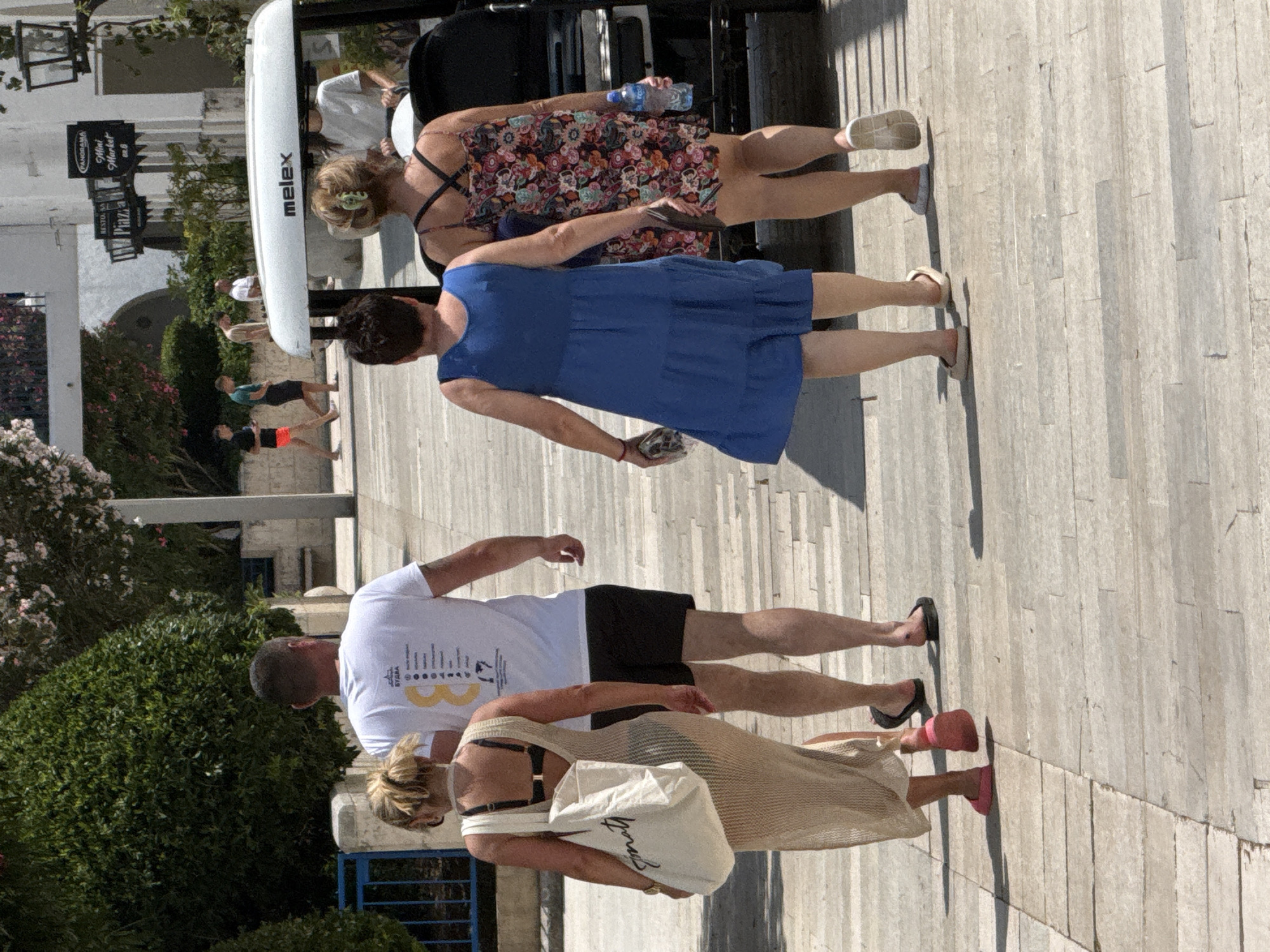 This is a rear-view, medium shot photograph taken outdoors on a sunny day in the city of Budva, Montenegro. Four individuals are walking away from the camera on a light-colored, paved walkway lined with trees and buildings.
In the foreground, two women are walking. The woman on the left is wearing a long, beige, knitted dress over a black bikini top and carrying a large, off-white tote bag. She is wearing pink flip-flops. The woman on the right is wearing a floral print sundress and is holding a plastic water bottle in her right hand. She is wearing off-white flats.
Behind the women, two men are walking. The man on the left is wearing a white t-shirt with a graphic print on the back and black shorts. The man on the right is wearing a light-colored t-shirt and dark shorts.
To the right of the group, a white golf cart with "melex" written on its roof is parked or slowly moving. In the background, there are buildings with signs, including "Panorama Mini Market" and "Resto Bar Plaza." Trees with green foliage and some pink flowers are visible, as is a blue railing. The overall scene depicts people in a public area, likely a resort or tourist district, in Budva.