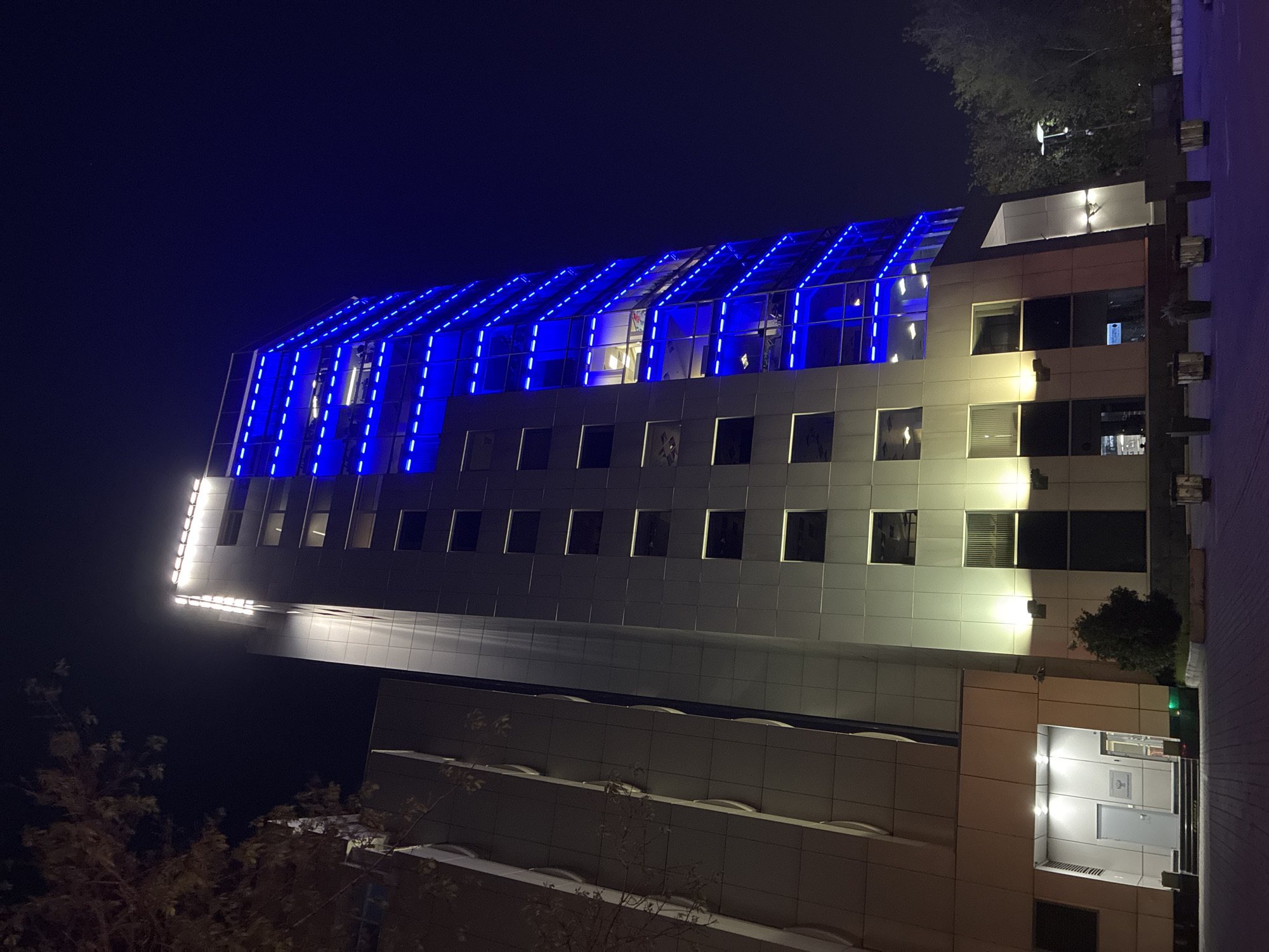A low-angle, full shot captures a multi-story building illuminated at night, likely in Moscow, Russia. The building's design features alternating bands of light and shadow, highlighting its architectural structure. The top of the building is distinguished by blue linear lighting that accentuates its geometric shape. Windows are visible, some reflecting internal lighting, while others appear dark, contributing to the contrast. Trees and other foliage are visible on the sides, adding to the urban landscape. The scene is cast in darkness, except for the artificial lighting that defines the building's features and surrounding elements. A walkway or road is partially visible, suggesting an accessible urban setting.
