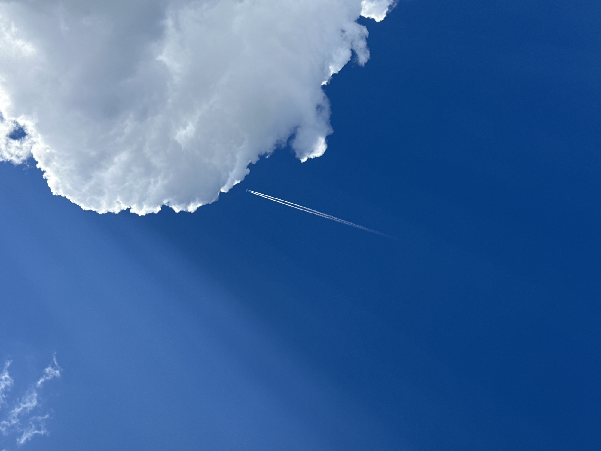 This is a vertical, medium shot photograph taken from a low angle looking up at a bright blue sky. A large, fluffy white cloud dominates the right side of the frame, with its edge casting a shadow across the sky from the upper left. A thin, white contrail from an airplane is visible diagonally across the lower left portion of the image, extending upwards and slightly to the right, disappearing behind the large cloud. The sky is clear and appears to be daytime. The image does not contain any visual information that allows for the identification of a specific location within the city of Estosadok, Russia.
