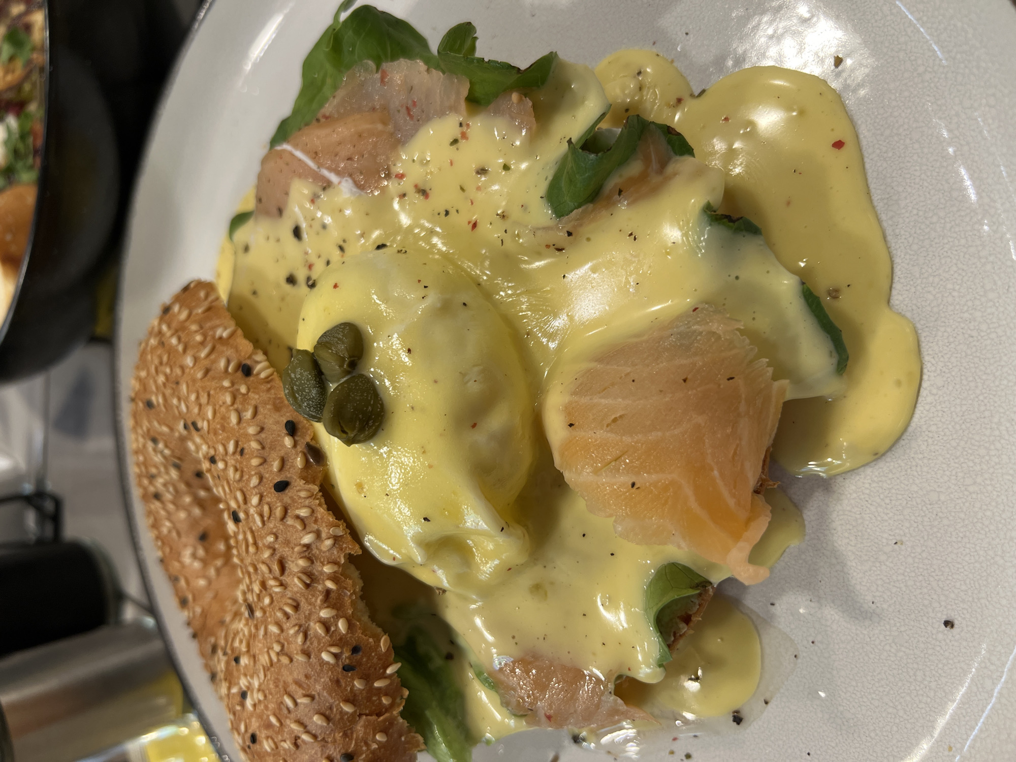 A close-up, overhead shot captures a plate of Eggs Benedict with salmon on a sesame bagel. The dish is liberally drizzled with Hollandaise sauce and sprinkled with pepper. Green leaves peek out from under the toppings. Capers sit atop the dish. The plate is white with a speckled grey rim. The location is Tbilisi, Georgia.