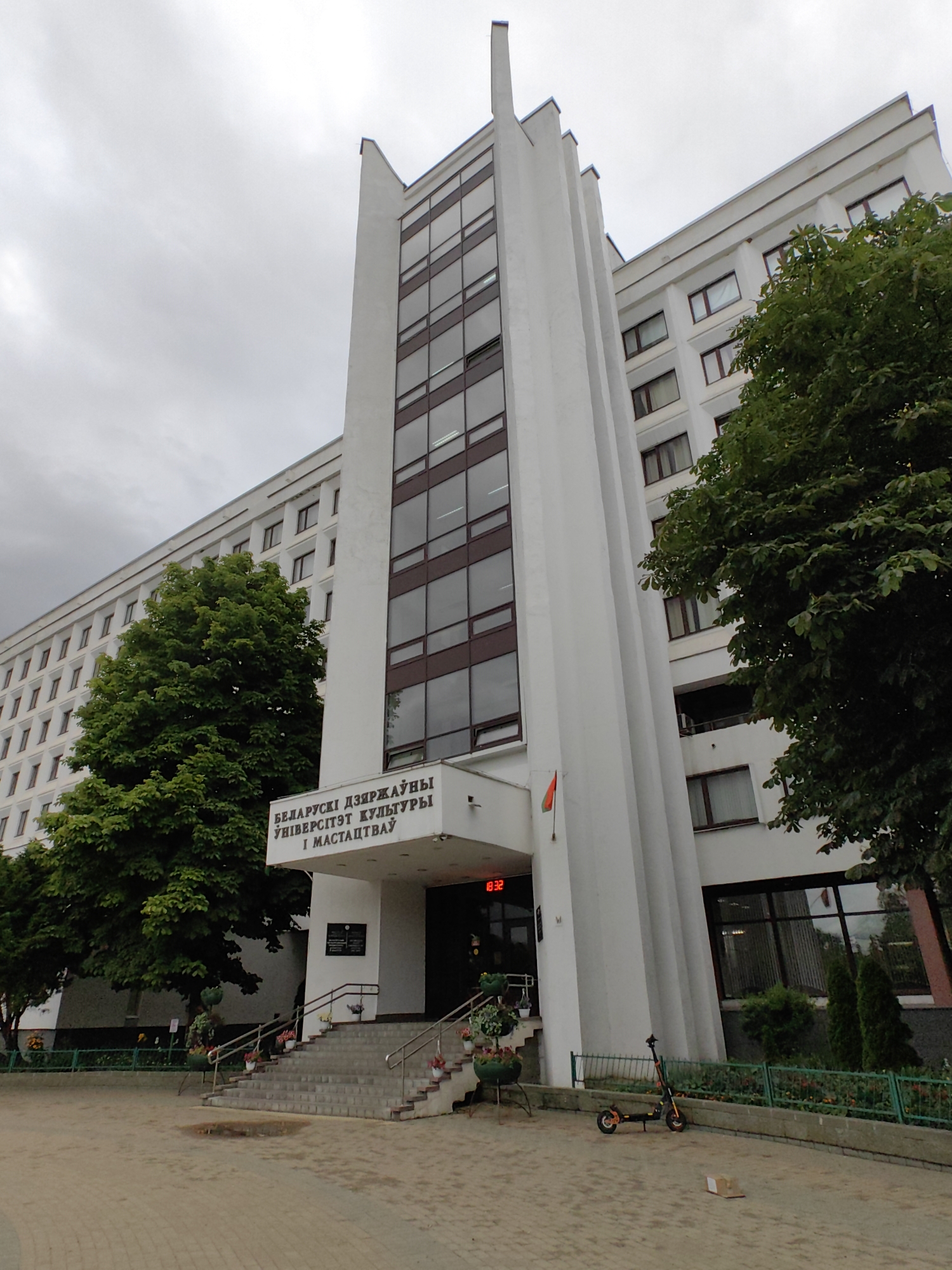 The image shows a wide shot of the Belarusian State University of Culture and Arts in Minsk, Belarus. The building is a large, white, multi-story structure with a prominent central tower featuring a grid of dark windows. The facade has a minimalist, Soviet-era architectural style. To the left of the central tower, a large, lush green tree is visible, partially obscuring the building. In front of the building, there is a wide set of stairs leading to the entrance. The steps are flanked by planters with colorful flowers. Above the entrance, text in Cyrillic reads "БЕЛАРУСКИ ДЗЯРЖАЎНЫ УНІВЕРСІТЭТ КУЛЬТУРЫ І МАСТАЦТВАЎ" (Belarusian State University of Culture and Arts). A small Belarusian flag hangs to the left of the entrance. To the right of the stairs, an electric scooter is parked, and a cardboard box lies on the paved ground. The sky is overcast, suggesting a cloudy day.