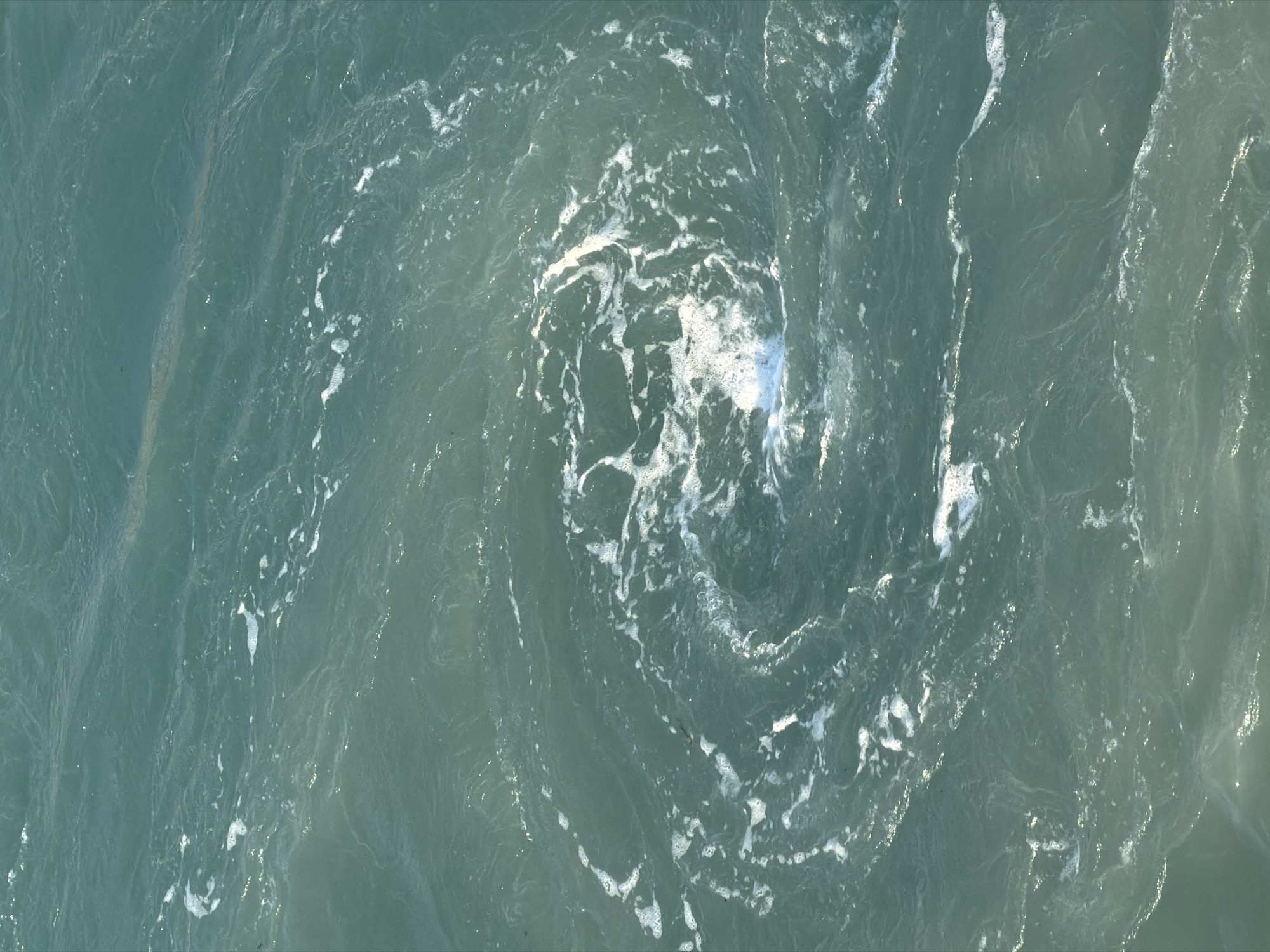 The image displays a close-up, overhead view of turquoise water with a swirling pattern and white foam. The water appears to be in motion, possibly a whirlpool or a strong current. There are no people or man-made objects visible in the frame. The texture of the water suggests it is a natural body of water, such as the sea or an ocean. The lighting indicates it is daytime, with sunlight reflecting off the surface of the water. Without further context, it is not possible to pinpoint the exact location within Zakynthos Municipal Unit, Greece, but it is likely a coastal area with active water movement.