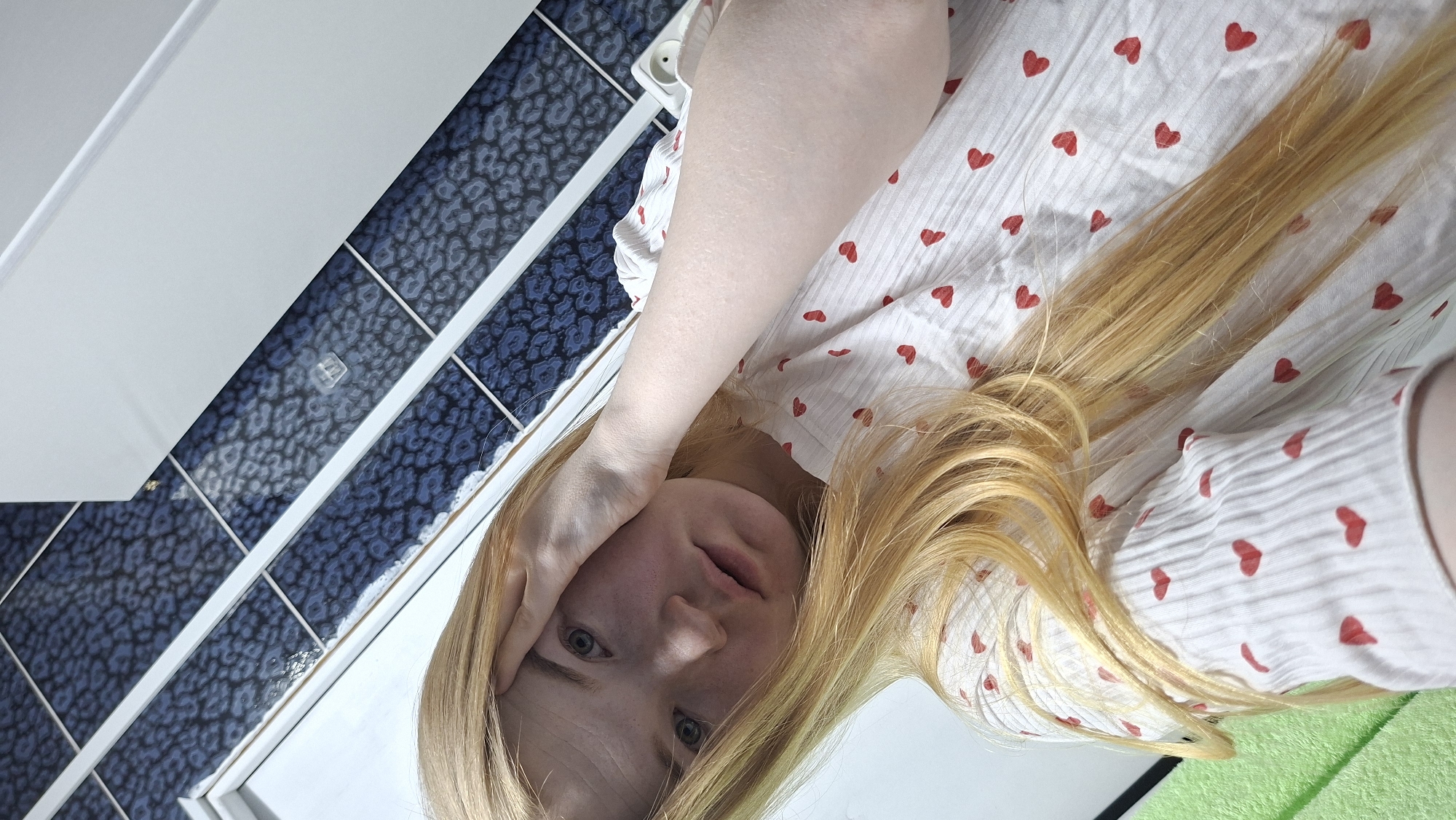 A fair-skinned person with blonde hair is angled towards the camera, their hand resting on their forehead. They wear a white shirt with red hearts. Behind them are white cabinetry and blue-patterned tiles. The location is identified as the city of Chaikovsky, Russia.