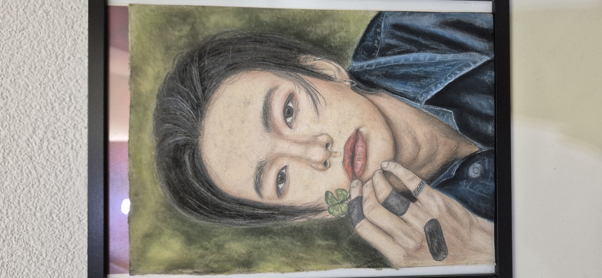 A framed portrait drawing of a person with dark hair and a denim jacket hangs on a white wall. The person in the drawing has pale skin, dark eyes, and red lips, and holds a four-leaf clover near their mouth. They wear rings on their fingers. The drawing is done in pastel or colored pencil and is framed in a simple black frame. The location is a building interior in Unknown, Unknown.