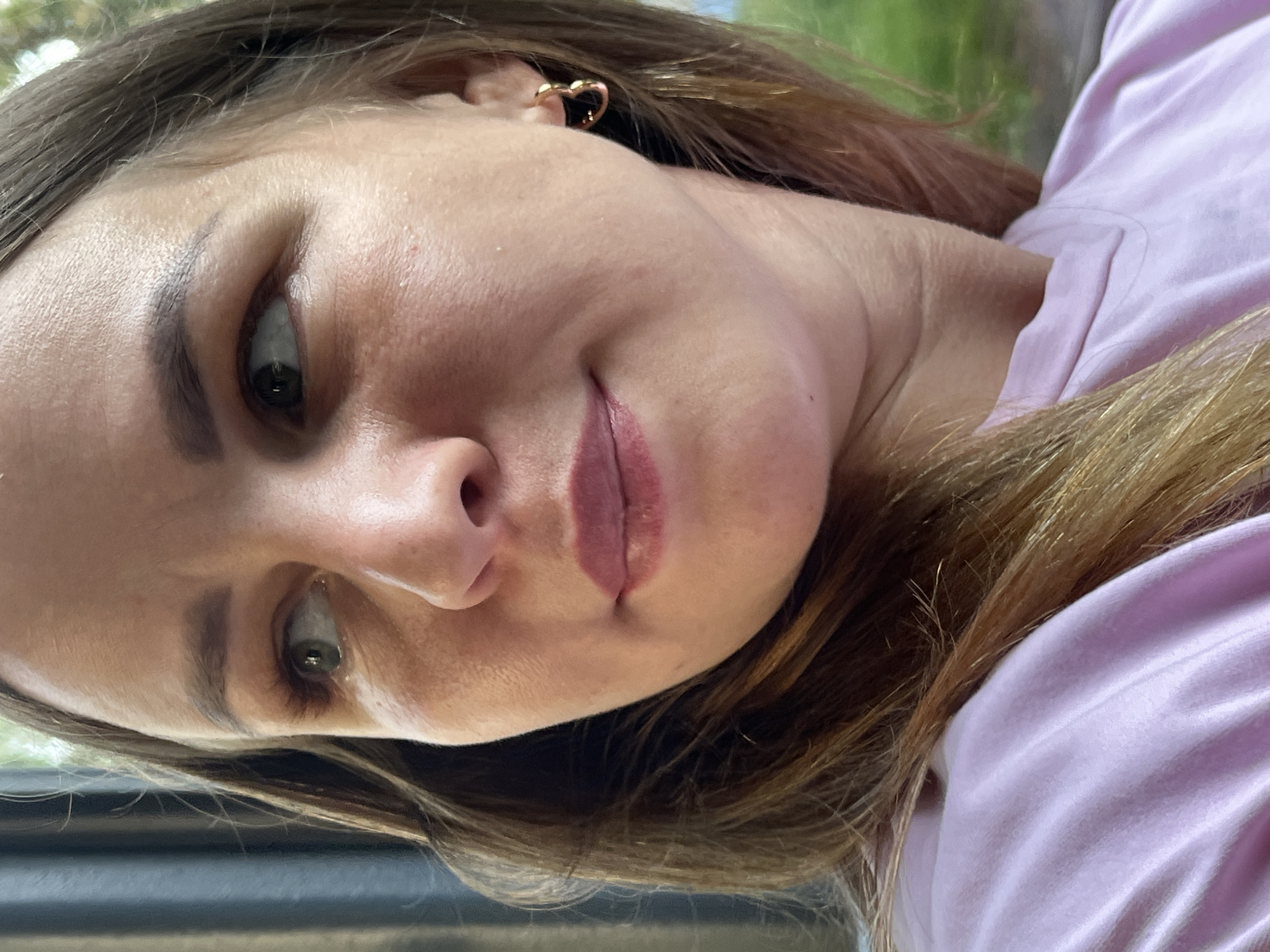 A woman with brown hair and green eyes is looking to the left of the camera. She is wearing a light purple t-shirt and a gold heart-shaped earring in her right ear. The image appears to be taken outdoors, with a blurry green background suggesting trees or foliage. The lighting suggests it is daytime. The location is unknown and cannot be determined from the image.
