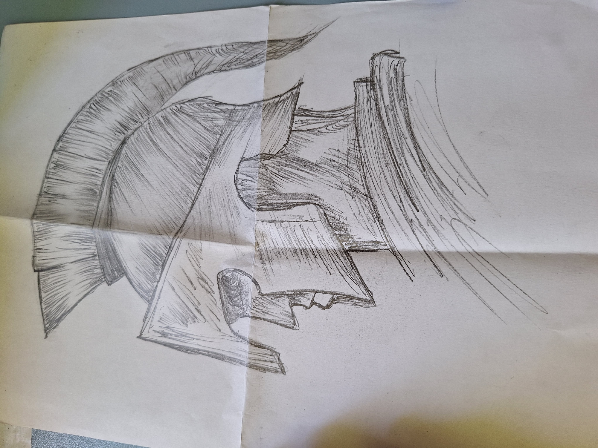 The image is a pencil sketch of a Spartan helmet in profile. The helmet is highly detailed, with lines representing the crest of feathers and the metal plating. The face of the warrior, or the visor of the helmet, is also depicted in profile, with a sharp nose and a hint of an eye. The sketch is on a piece of paper that appears to be folded, creating a crease across the middle. The background is plain, with no discernible environmental details. There is no information in the image that suggests a location in Varna, Bulgaria.