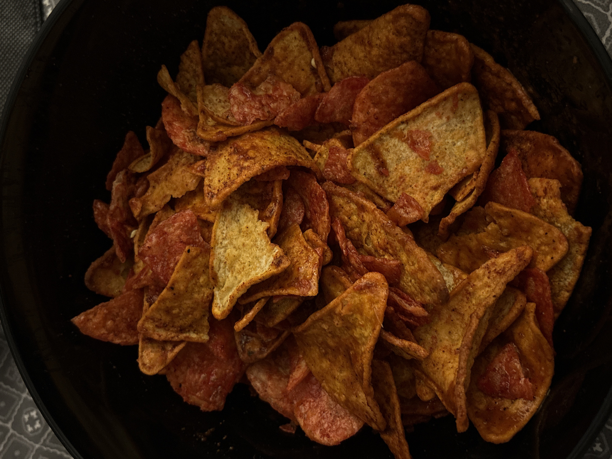 A close-up, high-angle shot shows a black bowl filled with numerous tortilla chips. Some chips appear golden brown with a seasoned surface, while others have a reddish tint, possibly due to a different type of flavoring or ingredient. The bowl sits on a surface that appears to have a patterned fabric or material. The setting or location within the city of San Bernardino Tlaxcalancingo, Mexico, is not identifiable from the provided image.