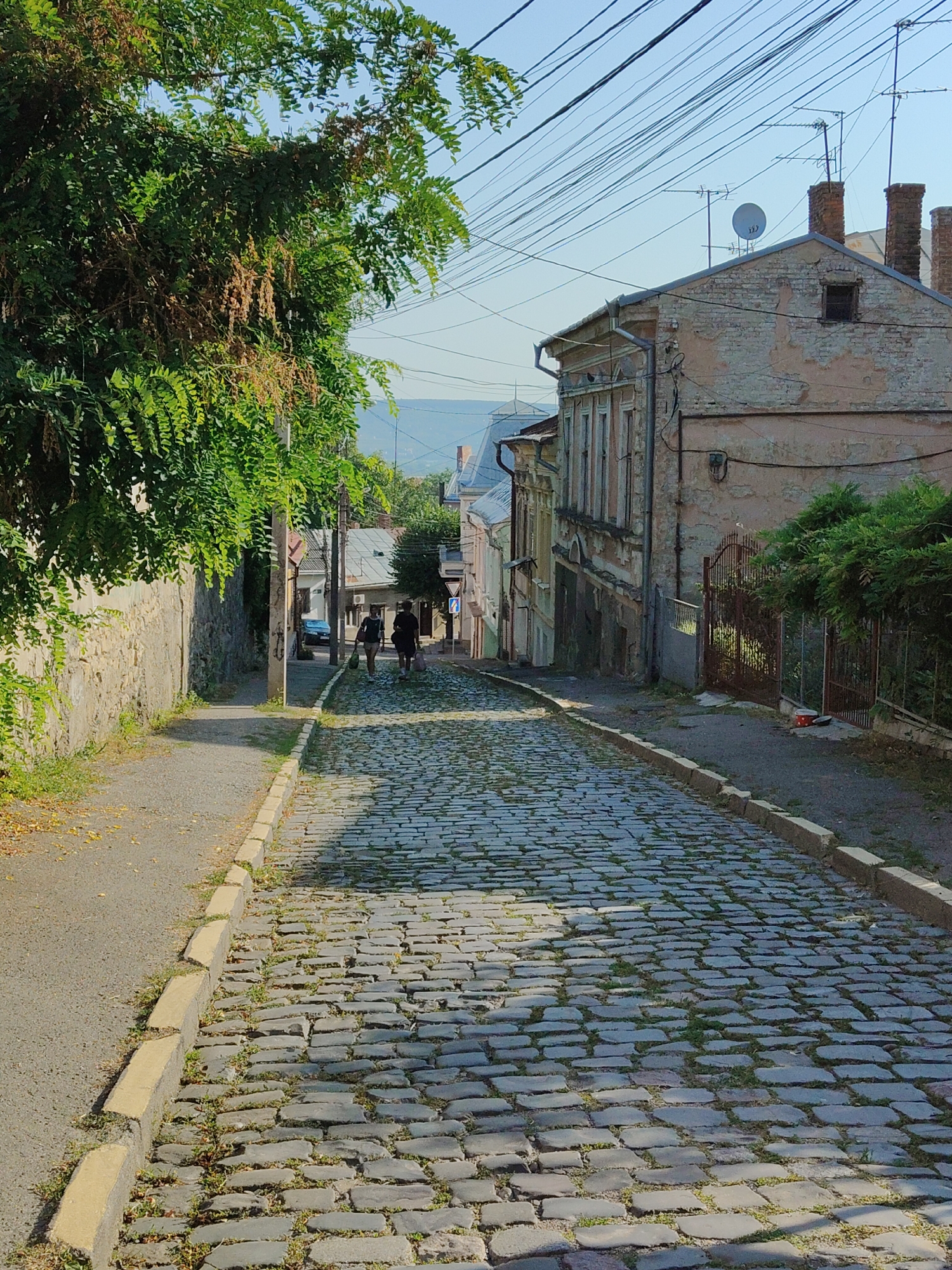 The image depicts a narrow, cobblestone street in Chernivtsi, Ukraine, on a clear day. The street slopes upwards and curves slightly to the right. On the left, a stone retaining wall is covered in lush green foliage. A paved sidewalk runs alongside the wall. On the right side of the street, old buildings with weathered facades line the road. One building in the foreground has exposed brickwork and a rusty metal fence. Several antennas and a satellite dish are visible on the rooftops. Two individuals, appearing to be walking away from the camera, are present on the cobblestone street. One is carrying a dark bag. The overall scene is characteristic of an older European city with a mix of natural elements and historic architecture.