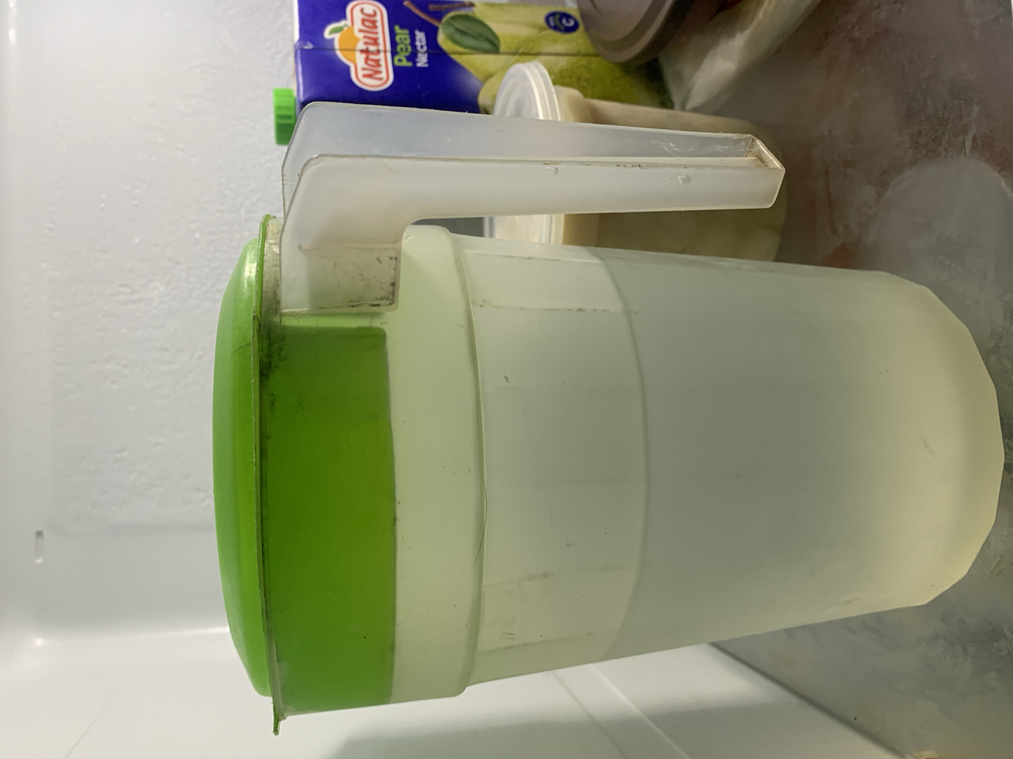 The image, taken in Santa Ana, Venezuela, displays the interior of a refrigerator, with the primary subject being a large, translucent plastic pitcher positioned horizontally in the frame. The pitcher features a bright green lid and a clear plastic handle. Dark discoloration, possibly mold or dirt, is visible along the rim where the green lid meets the pitcher's body, and also around the base of the handle. A light-colored liquid appears to be inside the pitcher.
In the background, a blue cardboard juice box is partially visible, oriented on its side. It is clearly labeled "Natulac Pear Nectar" with a pear graphic and an orange-colored brand logo. Another smaller, translucent plastic container with a white lid is partially seen behind the juice box. The interior walls of the refrigerator, white and textured, form the background of the scene. No specific activity is depicted; the image simply shows various items stored within a refrigerated space.
