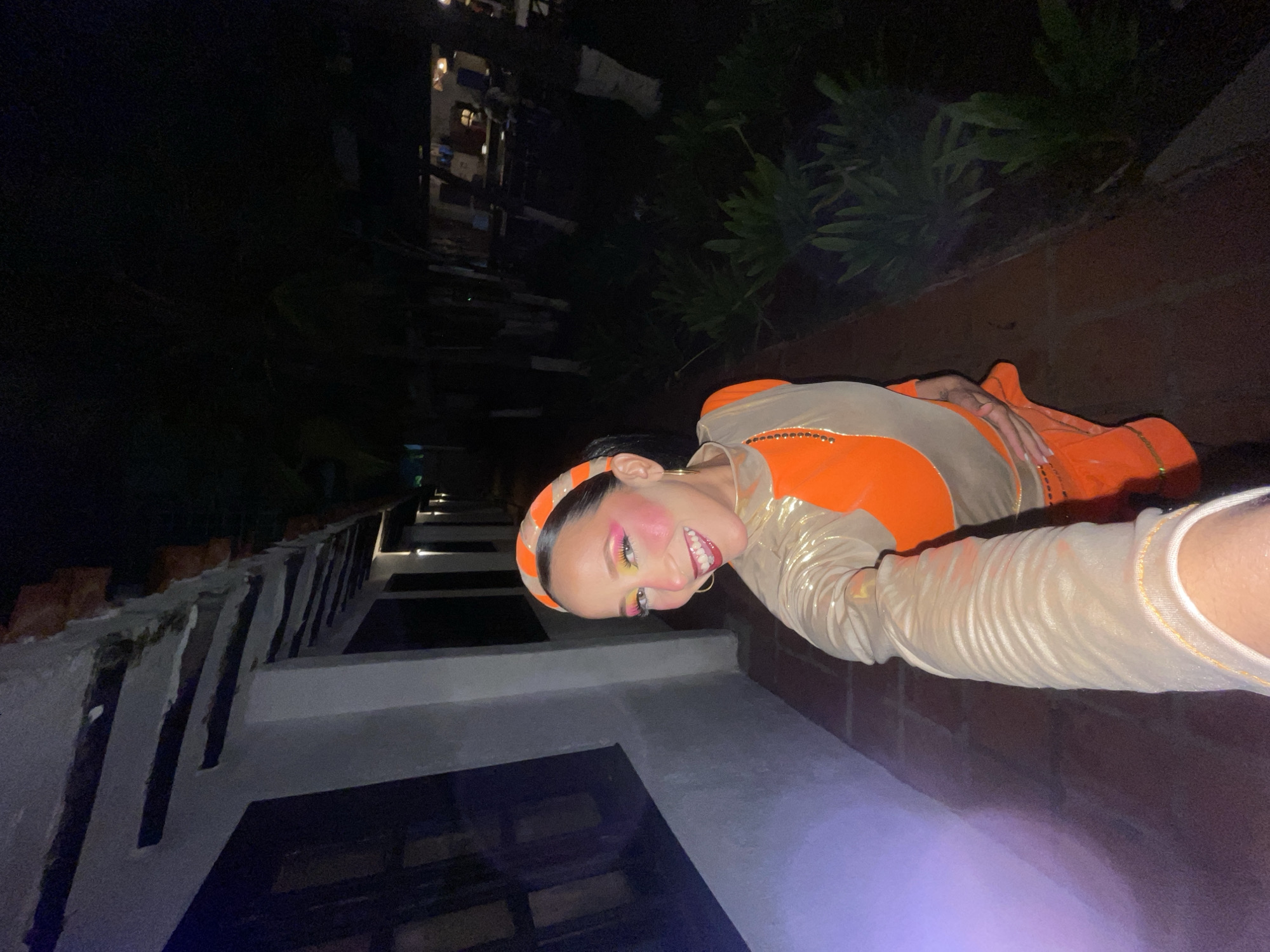 A woman in a brightly colored orange and gold outfit with dramatic makeup poses for a selfie at night. She is standing on a brick pathway in what appears to be an outdoor, possibly resort, setting with a white building to her left and lush greenery to her right. The building has dark-paned windows and exposed wooden beams supporting an overhang. Lights are visible along the building and further in the background, suggesting a commercial or hospitality establishment. The woman's outfit features a high neckline with gold trim and sheer, ruched sleeves. Her makeup includes bold eyeshadow, blush, and lipstick. The location is implied to be Porlamar, Venezuela.