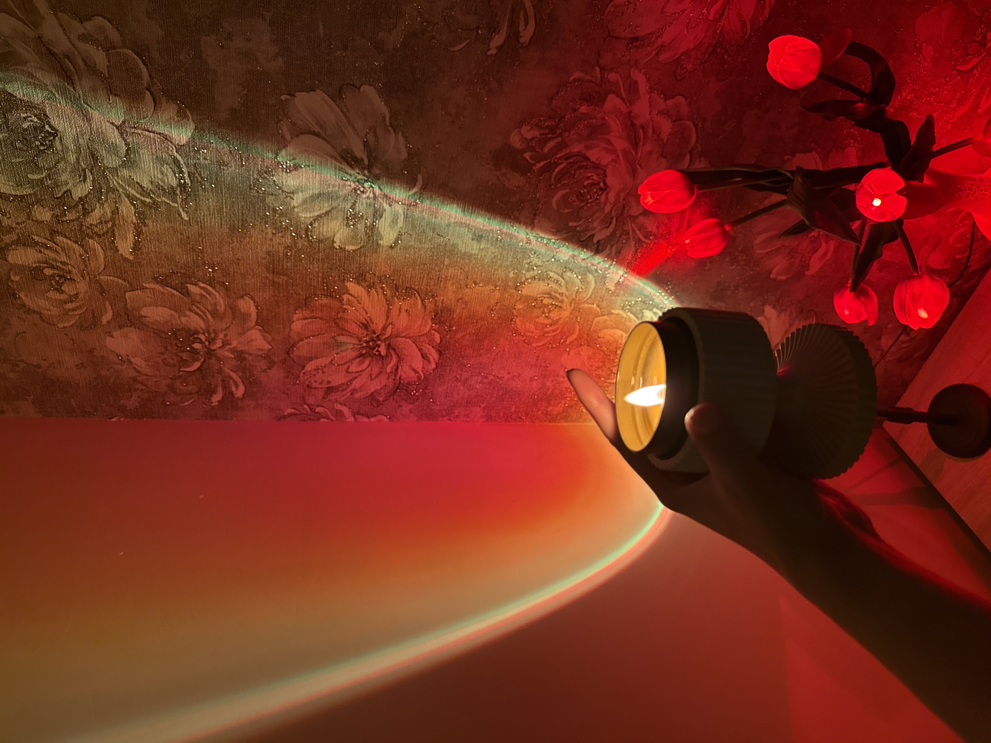 An indoor scene depicts a hand holding a lit candle. The candle is in a white container with a fluted base, and it emits a warm, yellow flame. The hand is positioned to the right of the candle. In the background, a wall with floral wallpaper is illuminated by a projected light that creates a colorful gradient of red, orange, and yellow. To the right of the candle, a vase holds several red tulip-shaped lights. The lighting in the scene is dramatic and atmospheric, with the projected light casting distinct shadows and highlights. The overall impression is one of a cozy and artistic interior setting.
