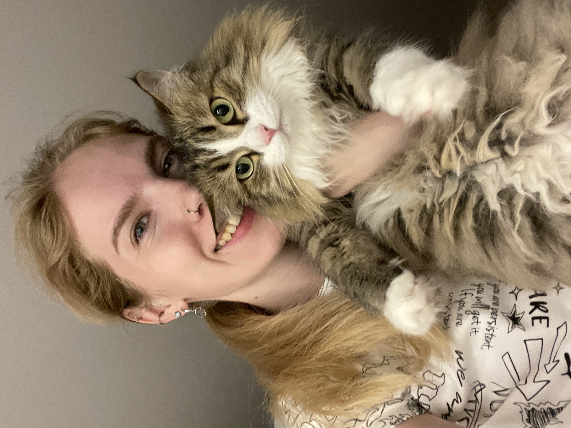 A close-up, vertically oriented indoor photograph features a young woman and a fluffy cat sharing a warm embrace. The woman, with light blonde hair, blue eyes, and a broad smile revealing her teeth, has multiple silver ear piercings and a nose ring. She is holding a large, long-haired tabby cat with distinctive green eyes, a white muzzle, and a mix of brown, grey, and white fur. The cat appears wide-eyed and alert, looking towards the camera.
The background is a plain, light grey wall, suggesting an intimate, uncluttered indoor setting in Izhevsk, Russia. The woman wears a light-colored top with black graphic designs and text. Visible phrases on her clothing include: "ARE," "you are persistent, you will get it," "If you are unsistent," and "we are." A star graphic and arrows are also part of the design. The image conveys a sense of happiness and affectionate bonding between the woman and her pet.