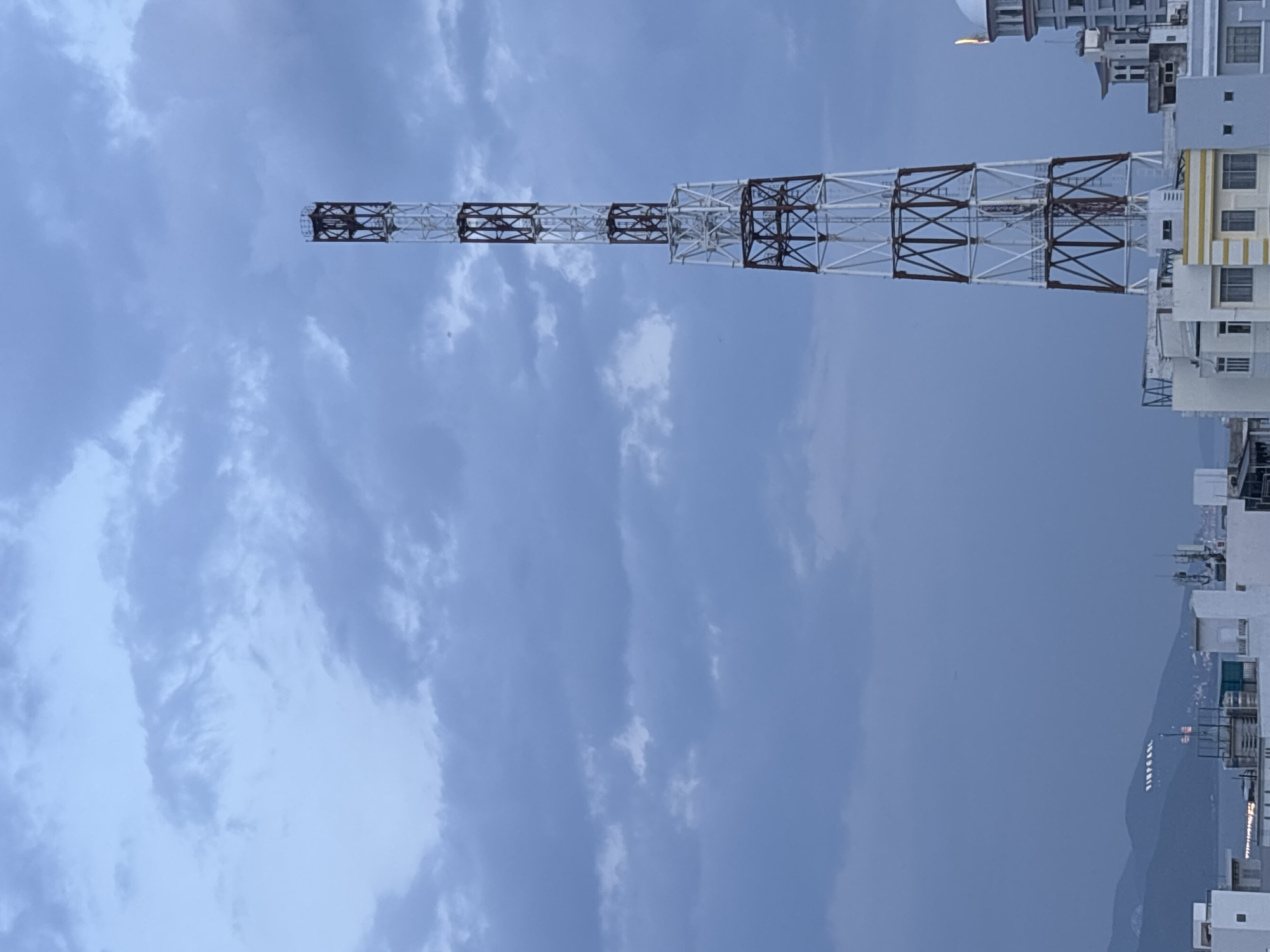 The image captures a tall, lattice-style tower with alternating white and reddish-brown sections, dominating the sky. The tower is positioned on the right side of the frame, with its peak extending high into a cloudy, blue-grey sky. In the foreground, the rooftops of several buildings are visible, some appearing modern with windows and balconies. To the far left, a range of hills or mountains can be seen, with some illuminated signage on their slopes, reading "VINPEARL" in capital letters. This signage, along with the mountainous landscape and the presence of buildings resembling a resort or urban development, strongly suggests the location is in or near Nha Trang, Vietnam, specifically in the Vinpearl complex area on Hon Tre island, or a viewpoint overlooking it. The overall lighting indicates it is either early morning or late evening, with diffused light and a sense of calm. No people are discernible in the image.