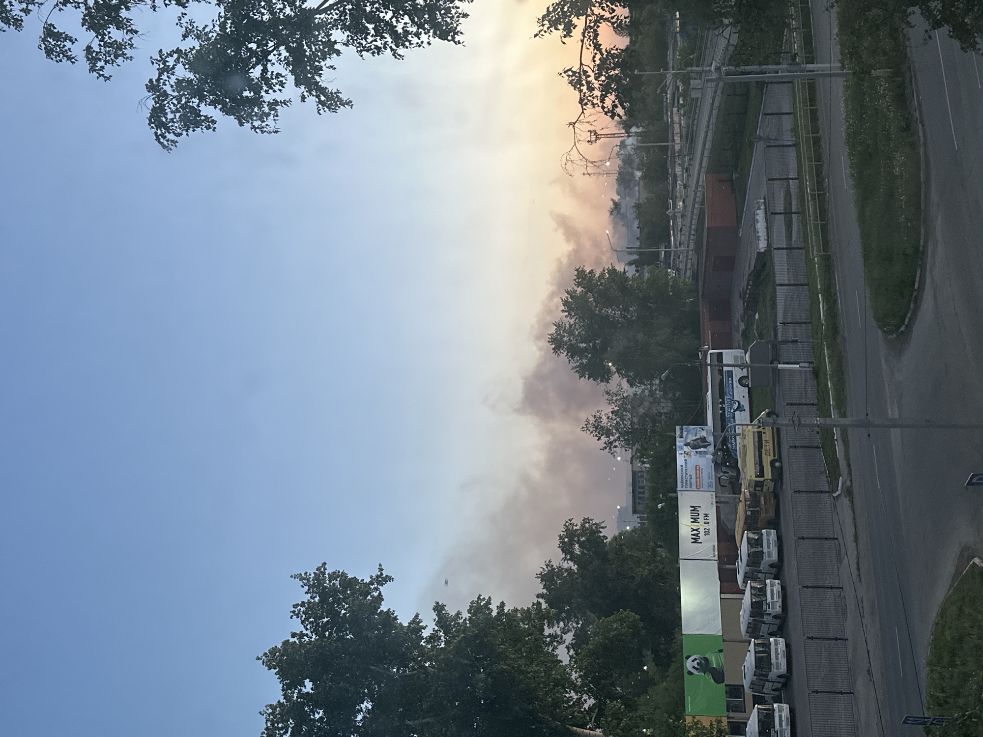 The image shows a view of Chaikovsky, Russia, with the sky dominating the upper half. The sky is blue on the left, transitioning to yellow and orange hues towards the right, possibly indicating sunrise or sunset. A layer of smoke or haze partially obscures the view of buildings and infrastructure.
In the lower half of the frame, rows of white vans and a yellow vehicle are parked along a street. Beyond them, a billboard is visible, partially obscured by trees. The billboard has an advertisement with the words "MAX MUM 102.0 FM", indicating a radio station.
Further in the background, obscured by haze, what appears to be industrial infrastructure and buildings are seen. In the foreground, trees frame the sky on either side of the shot. A road runs along the right side of the image. There are no visible people in the scene.