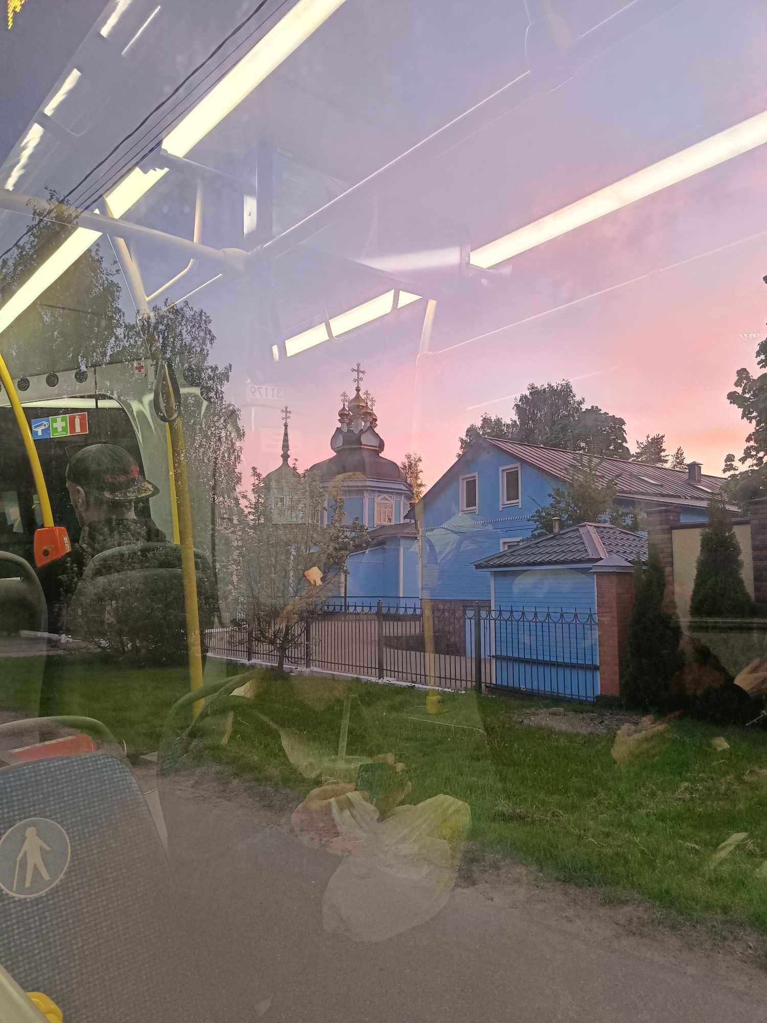The image shows a view from inside a bus in Saint Petersburg, Russia, looking out towards a blue building with a distinctive architectural style. This building is likely a church, recognizable by the ornate dome and cross. There is a person sitting on the bus, visible in the lower left of the frame. The outside environment consists of a grassy area, a fence, and trees. The sky is painted with a gradient of colors, suggesting sunrise or sunset. The overall impression is of a normal day on public transportation in Saint Petersburg, with a view of some of the city's cultural heritage.