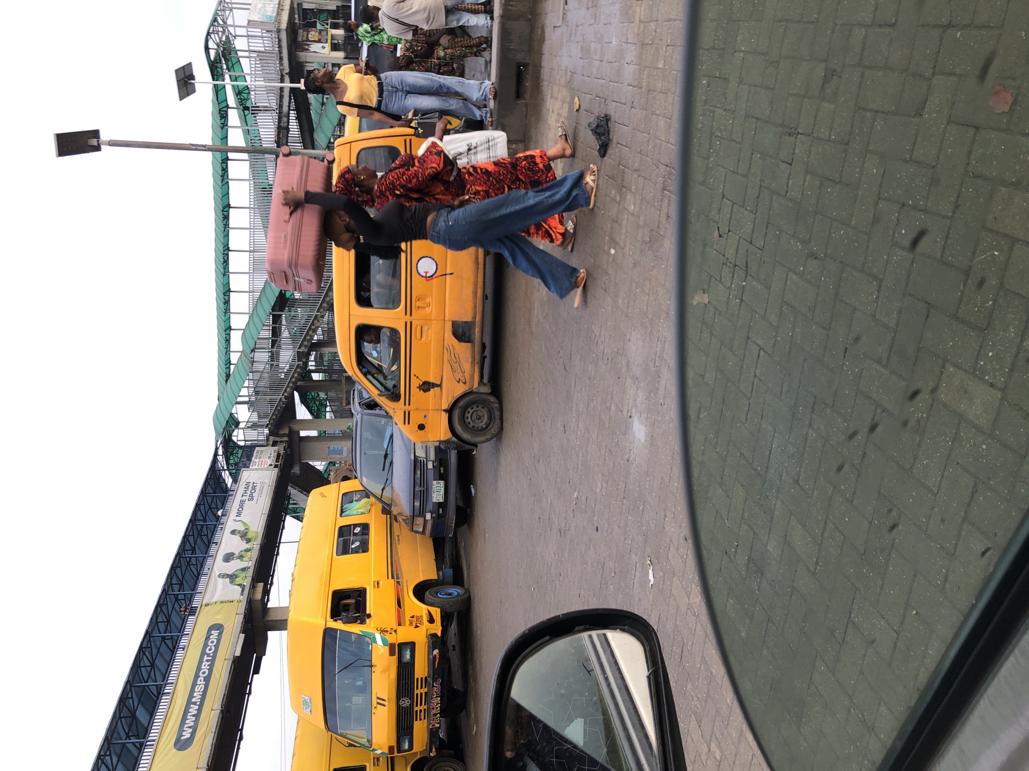 The image depicts a street scene in Ikeja, Nigeria, likely near a bus stop or transportation hub. In the foreground, reflections of the sky and pavement are visible in a car's side mirror and window. Behind the mirror, a yellow bus, a common mode of public transport in Lagos, is parked on a paved street. Another yellow bus is partially visible on the left. A dark-colored car is situated between the two yellow buses.
In the mid-ground, two women are walking. One woman is carrying a large pink suitcase on her head. She is wearing a black top and blue jeans. The other woman, beside her, is wearing a vibrant orange and red patterned dress and a headscarf. A third woman, standing to the right, is wearing a yellow t-shirt and blue jeans, looking towards the left. Further in the background, there's an elevated walkway with green roofing, typical of a pedestrian bridge, and various advertising banners. The scene appears to be a busy urban environment with elements of everyday life and transportation.