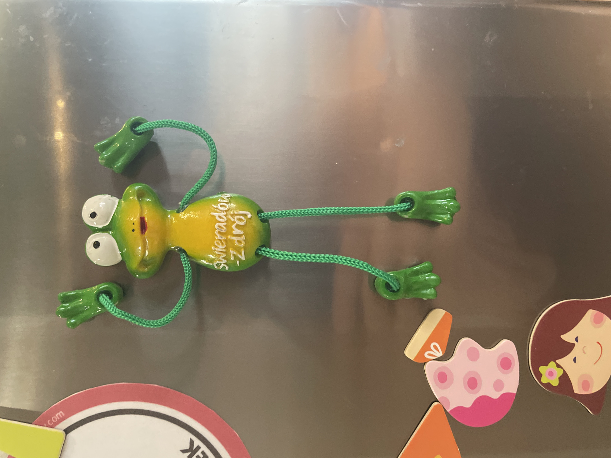 A magnetic decoration of a yellow and green frog is attached to a metallic surface, likely a refrigerator. The frog is posed with its green, braided arms and legs extended, its hands and feet resembling small, stylized paws. The frog's body is yellow with green accents on its back and head. White eyes with black pupils are visible on its head. White lettering on the frog's body reads "Świeradów-Zdrój," a place name. To the left of the frog, there are other magnets: a circular magnet with pink and white markings and a partial view of text that includes ".com" and "EK," and below that, a colorful puzzle piece magnet featuring a pink blob with white dots and a white outline. Further down, another magnet depicts the upper portion of a girl's face with brown hair, a flower behind her ear, and a smiling expression. The metallic surface reflects light in various areas, indicating it is a smooth, hard material. The background is otherwise unremarkable and appears to be the surface itself.