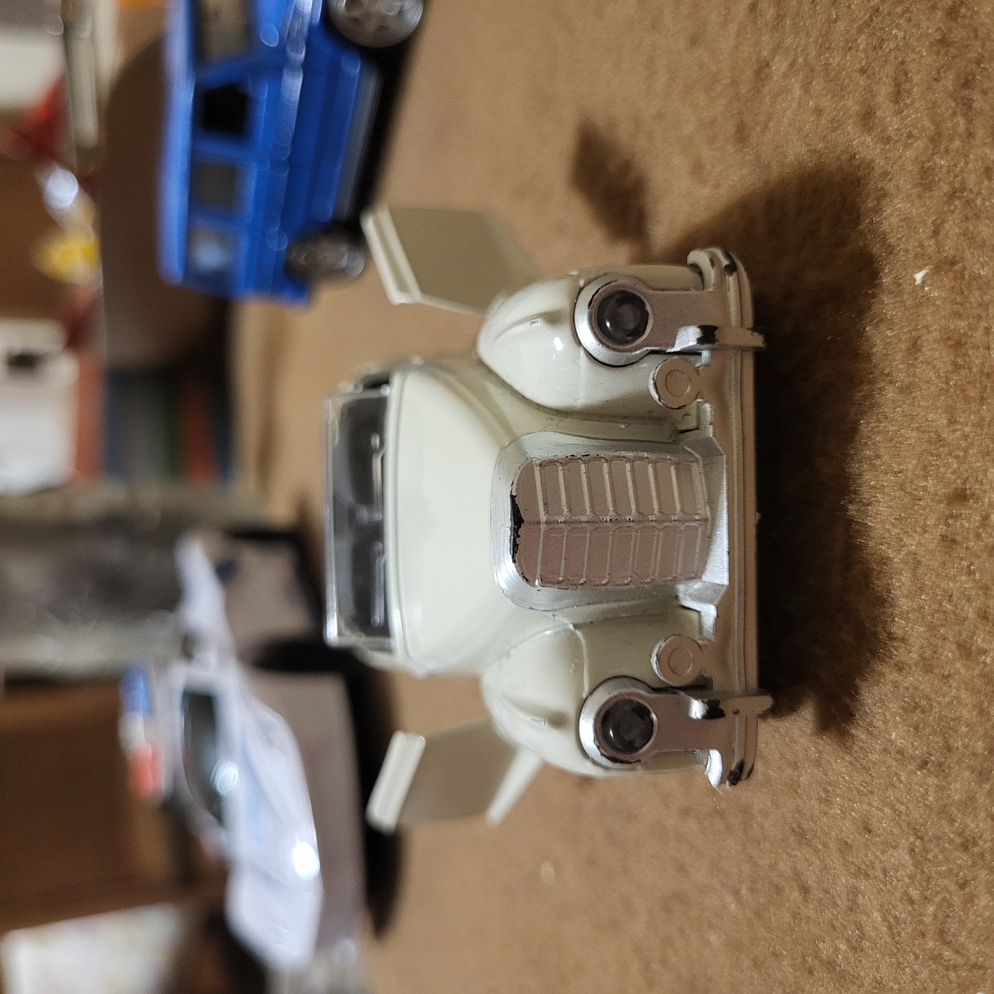 The image shows a close-up, top-down view of a vintage-style toy car, possibly a scale model, in a light cream color with silver accents. The car is positioned on a brown, textured surface, which appears to be fabric or carpeting. Visible details of the car include the headlights, radiator grille, fenders, and windscreen. Other toy vehicles, one blue and one white, are partially visible in the blurred background. There is no information that indicates that the image was taken in Yerevan, Armenia.
