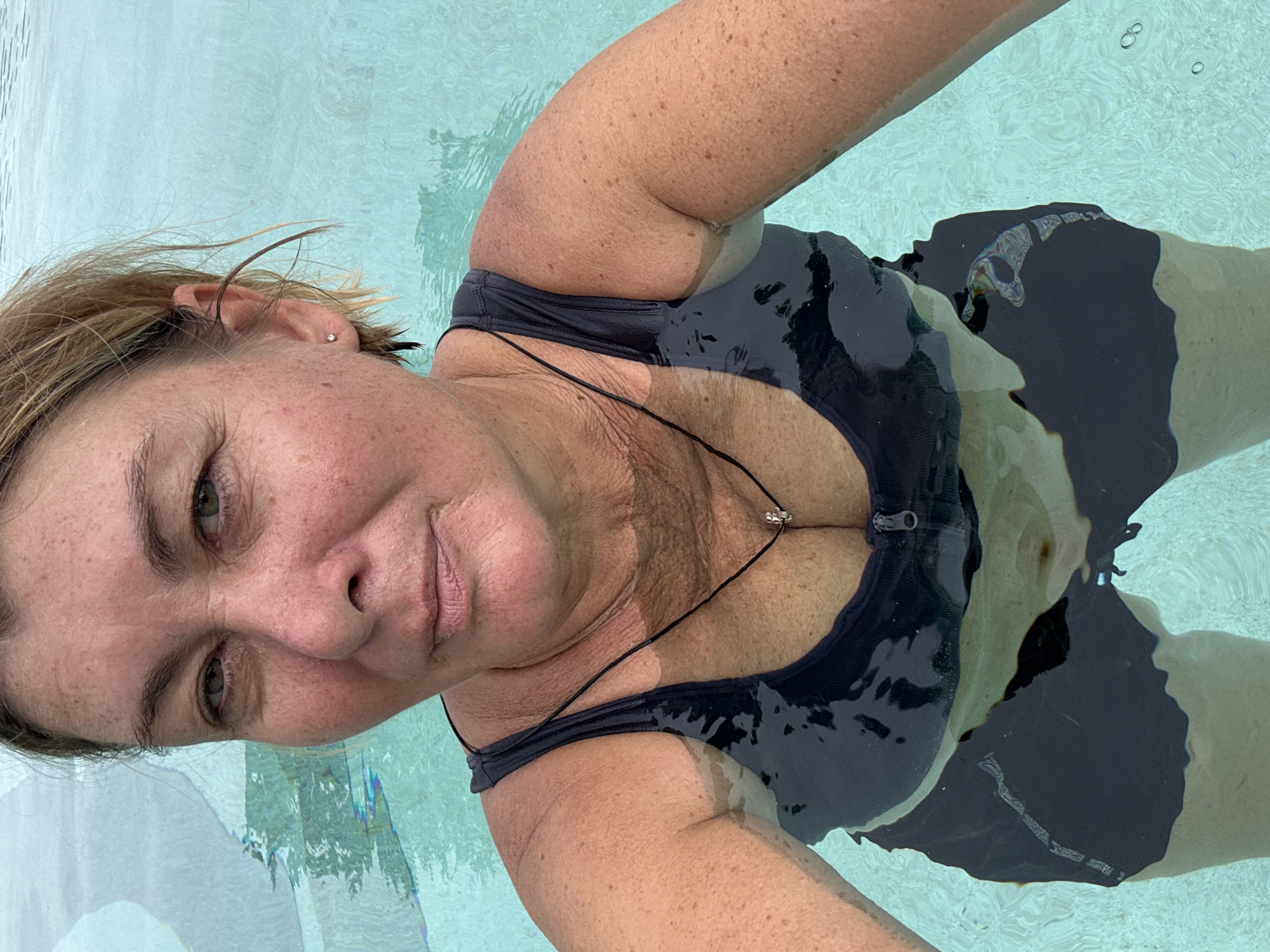 In the city of Ban Bang Rak, Thailand, a woman with blonde hair and freckled skin is pictured in a swimming pool. She is wearing a dark-colored swimsuit with a zipper front and a necklace with a small charm. The water is clear and blue, with light reflecting on the surface. The camera angle is close-up, capturing the woman's face and upper body.