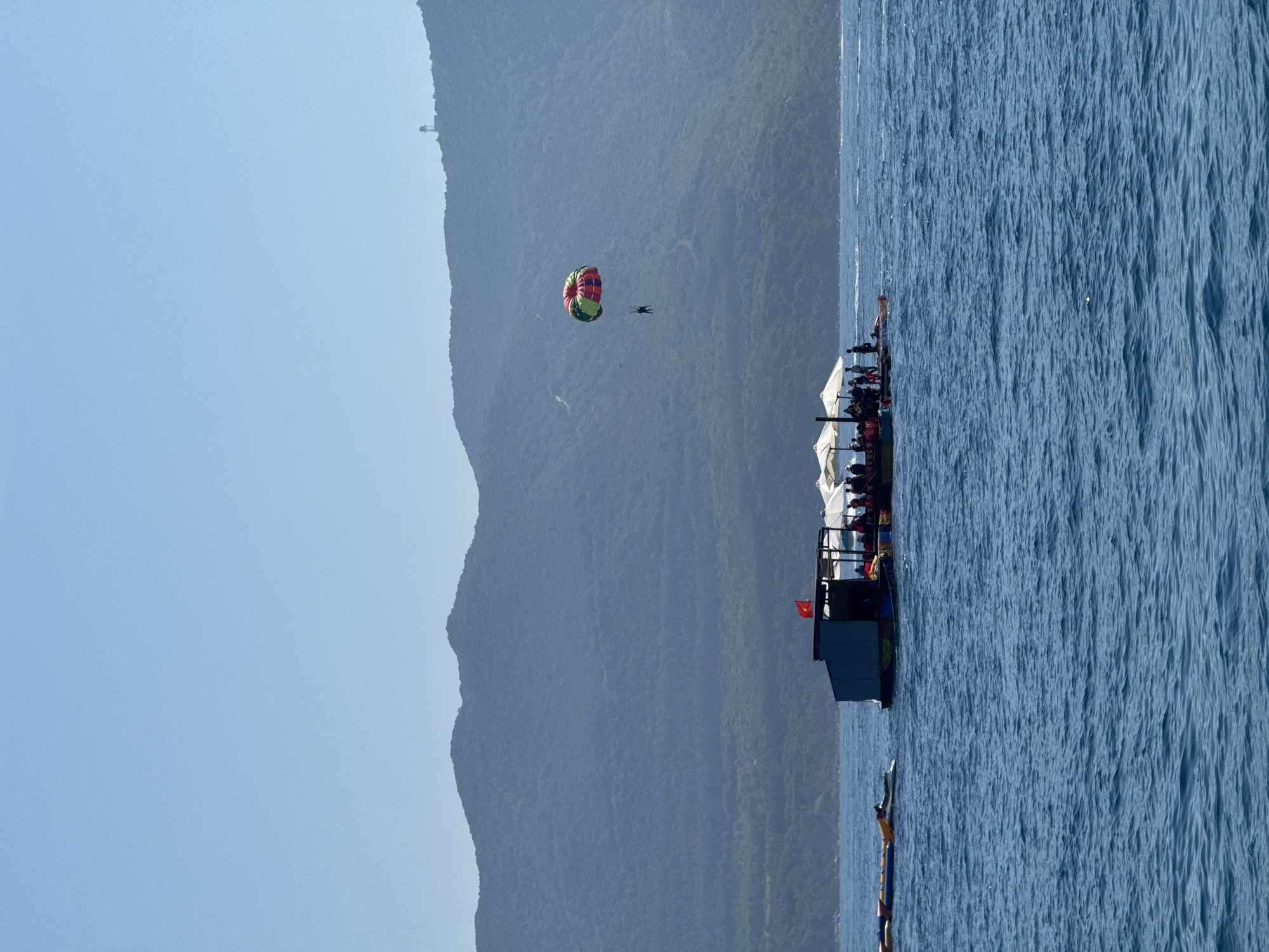 The image depicts a sunny day on the ocean off the coast of Nha Trang, Vietnam. In the foreground, the deep blue sea is calm with gentle ripples. In the middle ground, a raft-like structure is anchored, serving as a base for water activities. Several people are visible on the raft, some standing and others seated under white umbrellas. A Vietnamese flag flutters from a pole on the raft. To the left of the raft, a jet ski is partially visible. Further out in the water, a person is parasailing, suspended in the air by a colorful parachute against the backdrop of a large, verdant mountain range that dominates the horizon. The mountain slopes are covered in dense vegetation, and a small, distant structure, possibly a lighthouse or a tower, can be seen on the crest of the mountain. The sky is a clear, pale blue.
