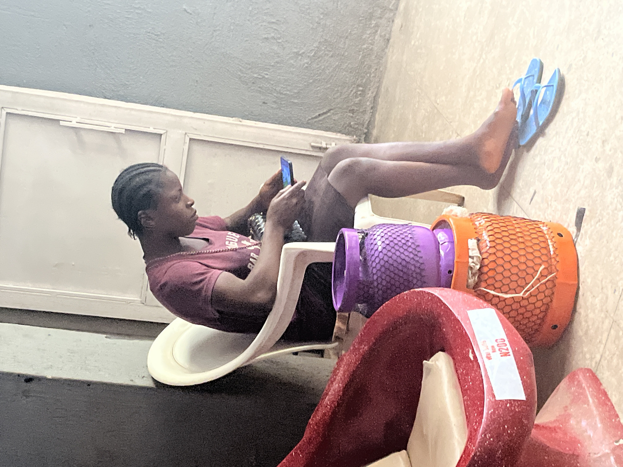 A young person sits in a white plastic chair, holding a smartphone in their hands, in what appears to be an outdoor or semi-outdoor area. The person is wearing a maroon t-shirt and dark shorts, with their hair braided. To the left of the chair is a large, red, curved object, possibly a decorative item or part of a structure, with a white sticker displaying "N200." Next to this red object are two stacked cylindrical items, the top one being purple with a textured surface, and the bottom one being orange and also textured. In front of the chair, on the floor, are a pair of blue flip-flops. The background consists of a light-colored door and a textured gray wall. The overall environment suggests a casual setting, potentially in Jos, Nigeria, given the context provided.