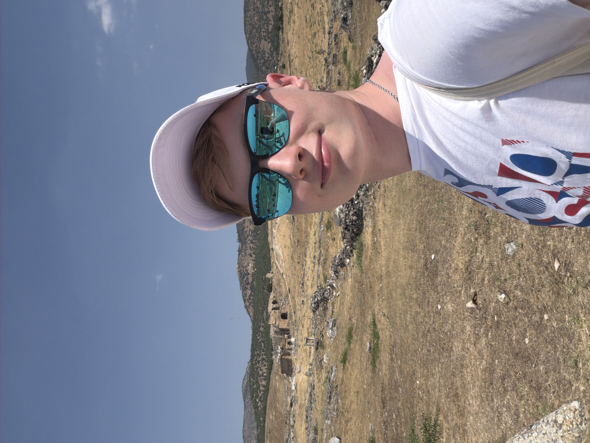 A young male with fair skin and blonde hair is taking a selfie outdoors in Pamukkale, Turkey. He is wearing a white baseball cap, a white t-shirt with a red, white, and blue graphic design, and a silver chain necklace. He has on blue mirrored sunglasses that reflect a scene of ancient ruins and other people. The background shows a dry, arid landscape with scattered rocks and the remnants of ancient structures, characteristic of the Hierapolis archaeological site in Pamukkale. Rolling hills covered in sparse vegetation and trees form the distant background under a clear blue sky. The overall scene is brightly lit by sunlight, suggesting it is a warm, sunny day.