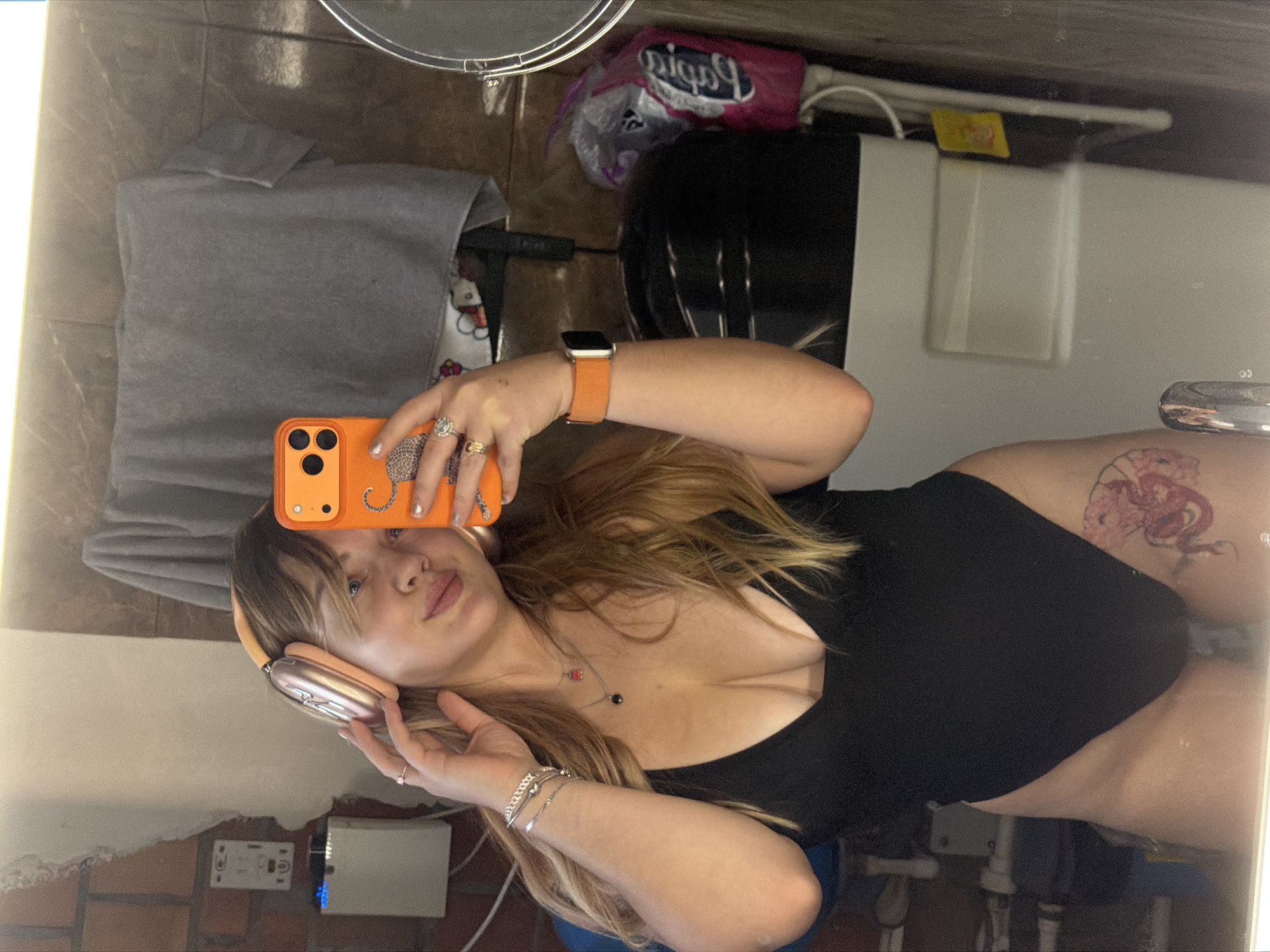 This is a mirror selfie capturing a young woman indoors. She has long, wavy blonde or light brown hair that cascades around her shoulders and chest. She is wearing a black top, possibly a swimsuit or bodysuit, revealing some cleavage. On her head are light peach or rose gold over-ear headphones. Her expression is neutral to slightly smiling.
She holds an orange smartphone with three visible camera lenses in her left hand, which has several rings: two on the ring finger, one on the pinky, and one on the index finger. On her left wrist, she wears an orange smartwatch. Her right wrist is adorned with multiple silver-toned bracelets. A prominent tattoo depicting a pink or red creature, possibly a dragon or sea monster, is visible on her right hip or upper thigh.
The reflection behind her shows various household items. To the upper left, a folded grey fabric, resembling a towel or blanket, is visible next to a white item with a Hello Kitty design. Above her left arm, a pink plastic bag with mirrored text that appears to read "Pigia" or similar is seen, alongside a dark object and wires. Further to the right, a white rectangular appliance or device is reflected. The bottom portion of the reflection shows a textured wall that resembles brickwork, with what looks like an electrical outlet and another small white device with a blue light. The lighting suggests an indoor setting, likely with artificial light. No specific location cues are discernible within the image itself beyond it being an interior space.