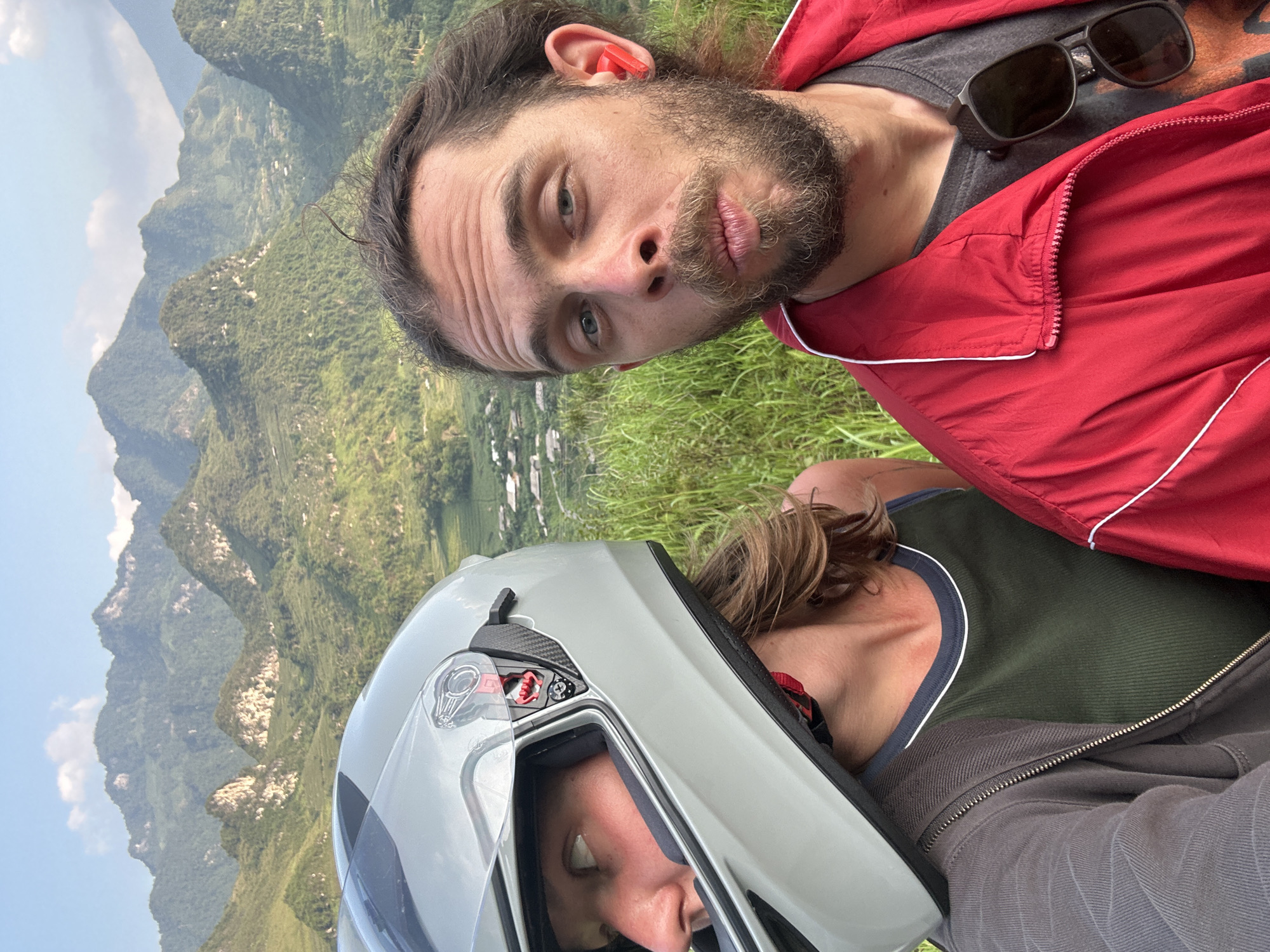 Two people are in the image, positioned against a backdrop of mountains and greenery. One person is wearing a red jacket and sunglasses resting on their chest. They have a beard and long hair. The other person is wearing a helmet, obscuring much of their face. The location is identifiable as the rural area of Du Già Commune, Vietnam, based on the distinctive mountain topography and vegetation.