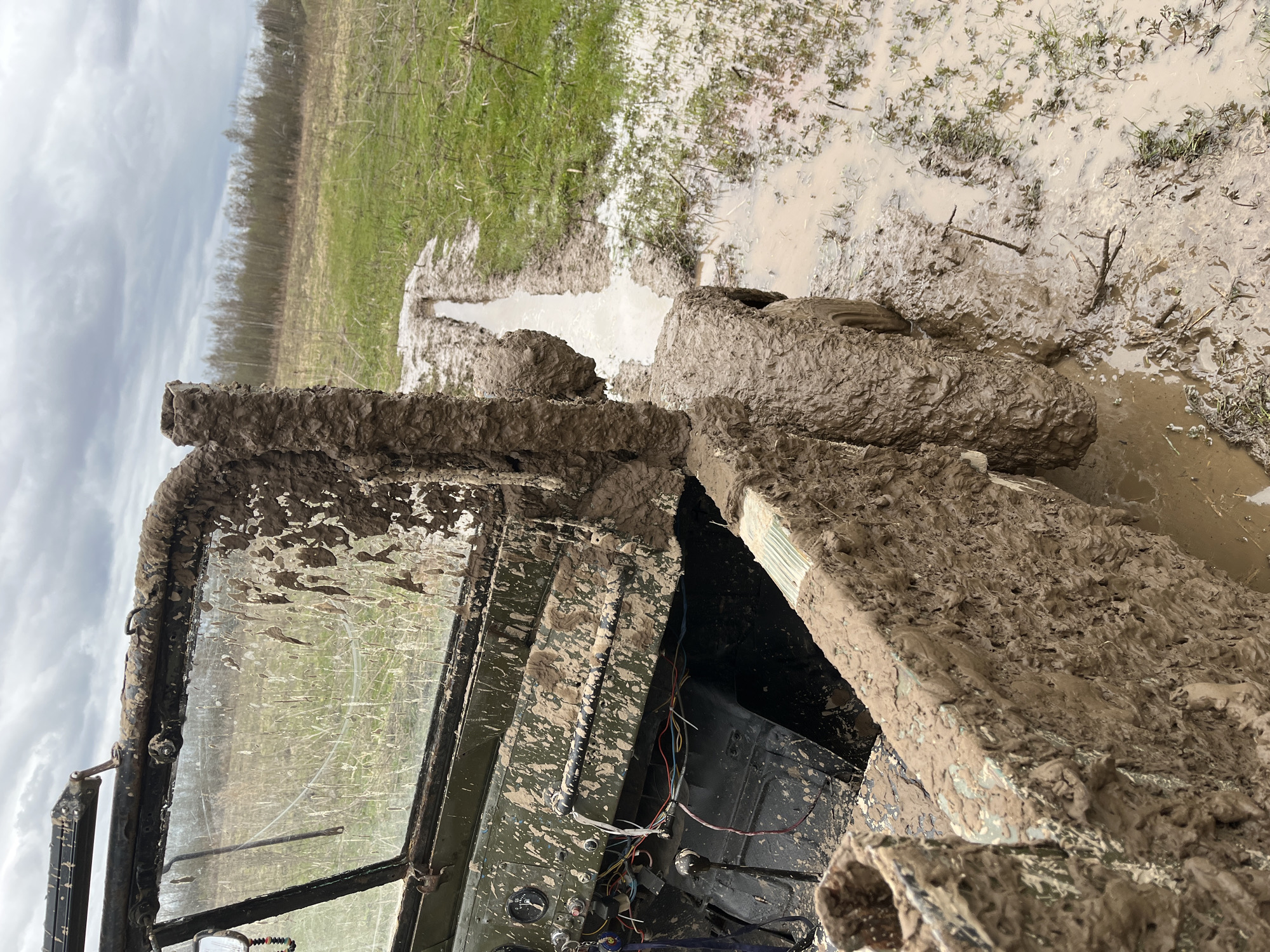 A mud-covered vehicle is mired in a muddy area, possibly near a field with some grass, in an area near Раздолье, Russia. The vehicle's windshield is splattered with mud, and the interior appears damaged with exposed wires. The wheels are deeply embedded in the mud, suggesting the vehicle is stuck. The surrounding landscape is flat and wet, with a narrow path of muddy water extending into the field. The sky is overcast, indicating recent or ongoing rainfall.
