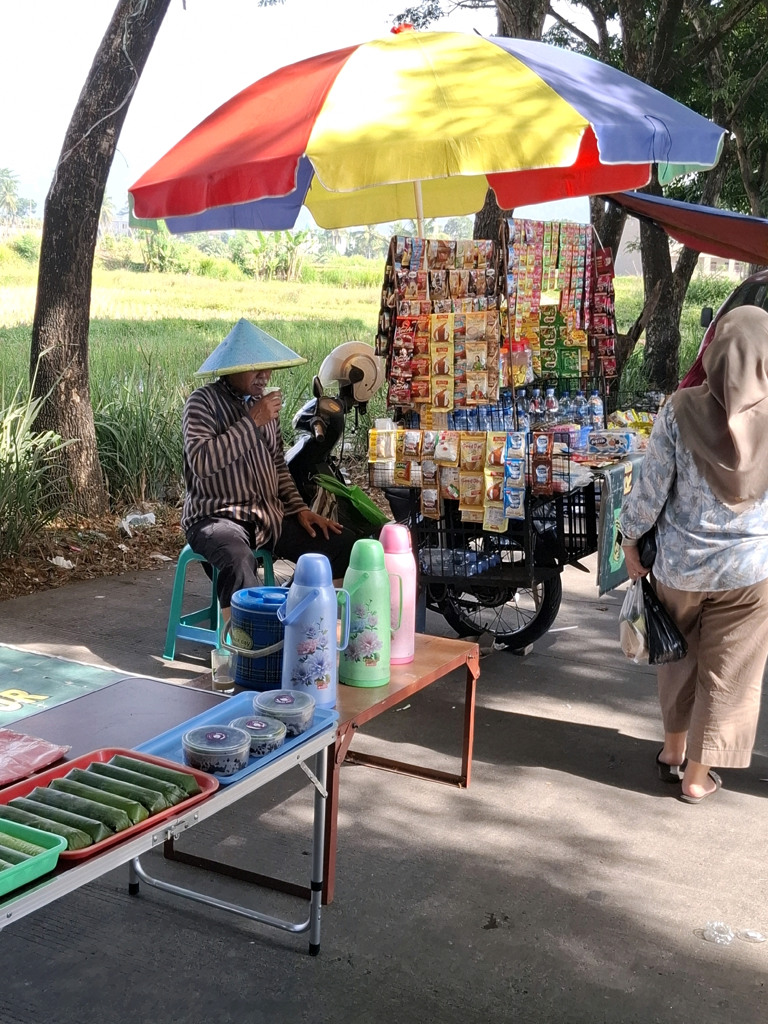 In Karangtengah, Indonesia, a man wearing a conical hat and striped shirt is seated on a blue stool, drinking from a cup. He is next to a motorcycle and a cart selling various packaged food items and drinks. A colorful umbrella shades the cart. In the foreground, a table displays food items wrapped in banana leaves, along with containers and thermoses. A woman wearing a hijab and brown pants walks away from the cart, carrying plastic bags. The scene takes place outdoors, with a background of green vegetation and trees, suggesting a roadside setting.