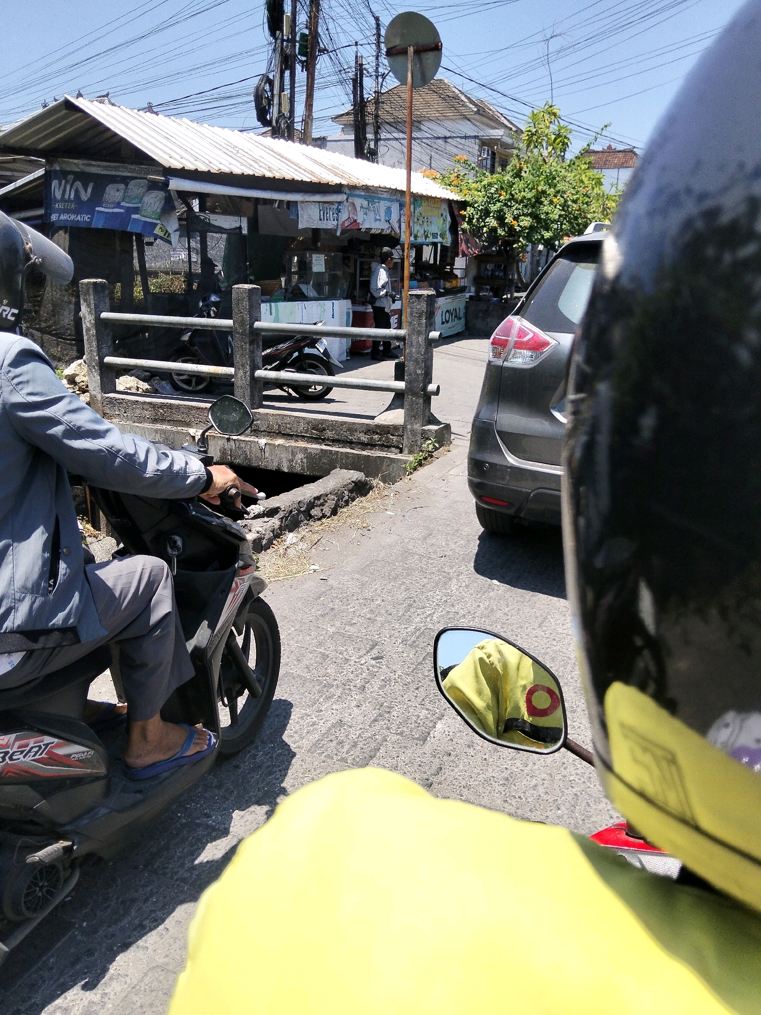 The image captures a street scene in Denpasar, Indonesia, likely during daytime due to the bright sunlight and clear blue sky. The perspective is from a person riding a motorcycle, showing their hand on the handlebar and their yellow jacket reflected in the side mirror. In the foreground, the rider's left leg and blue flip-flops are visible, resting on the motorcycle. The motorcycle itself is dark-colored with red accents.
To the left of the motorcycle, a shop with a white corrugated metal roof is visible. A banner on the shop displays the word "VIN" in large white letters and "KRETEK" and "SET AROMATIC" in smaller text. Several other signs and advertisements are present on the shop front. A metal railing with concrete posts separates the shop from the road. Behind the railing, more motorcycles are parked.
In the middle ground, a dark gray SUV is stopped on the road, its rear visible. The road appears to be asphalt. Further in the background, trees with green foliage and some orange flowers can be seen, along with buildings and power lines crisscrossing the sky. A round traffic sign on a pole is also present. The overall impression is that of a busy street in a tropical urban environment.