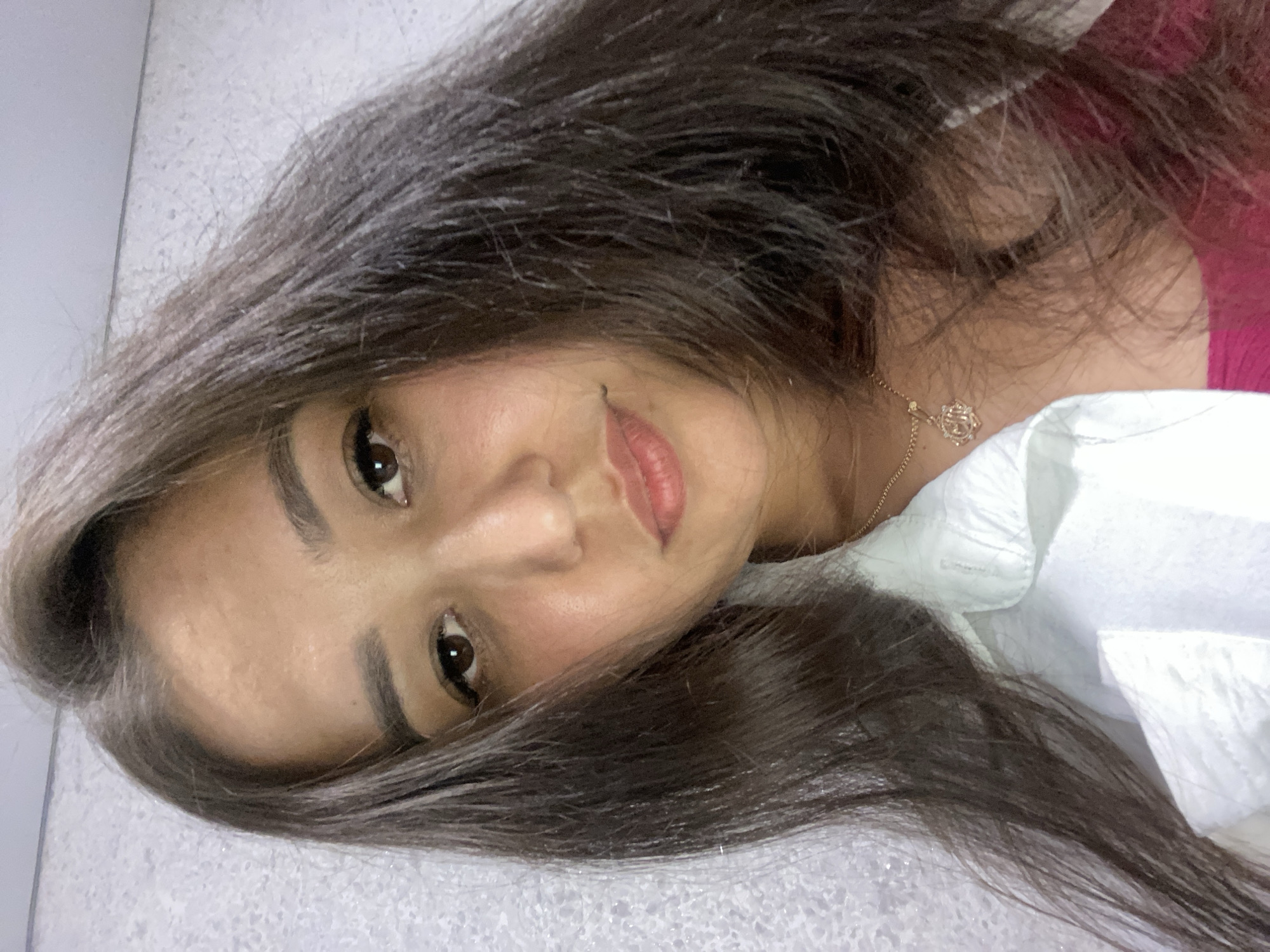 A young woman is shown in a close-up selfie, with her head tilted slightly. She has dark brown hair that falls around her face and shoulders. She is wearing a white collared shirt and a maroon top underneath, with a delicate gold necklace featuring a circular pendant. The background is a textured, light-colored wall. The image was taken in Ufa, Russia, in an indoor setting.