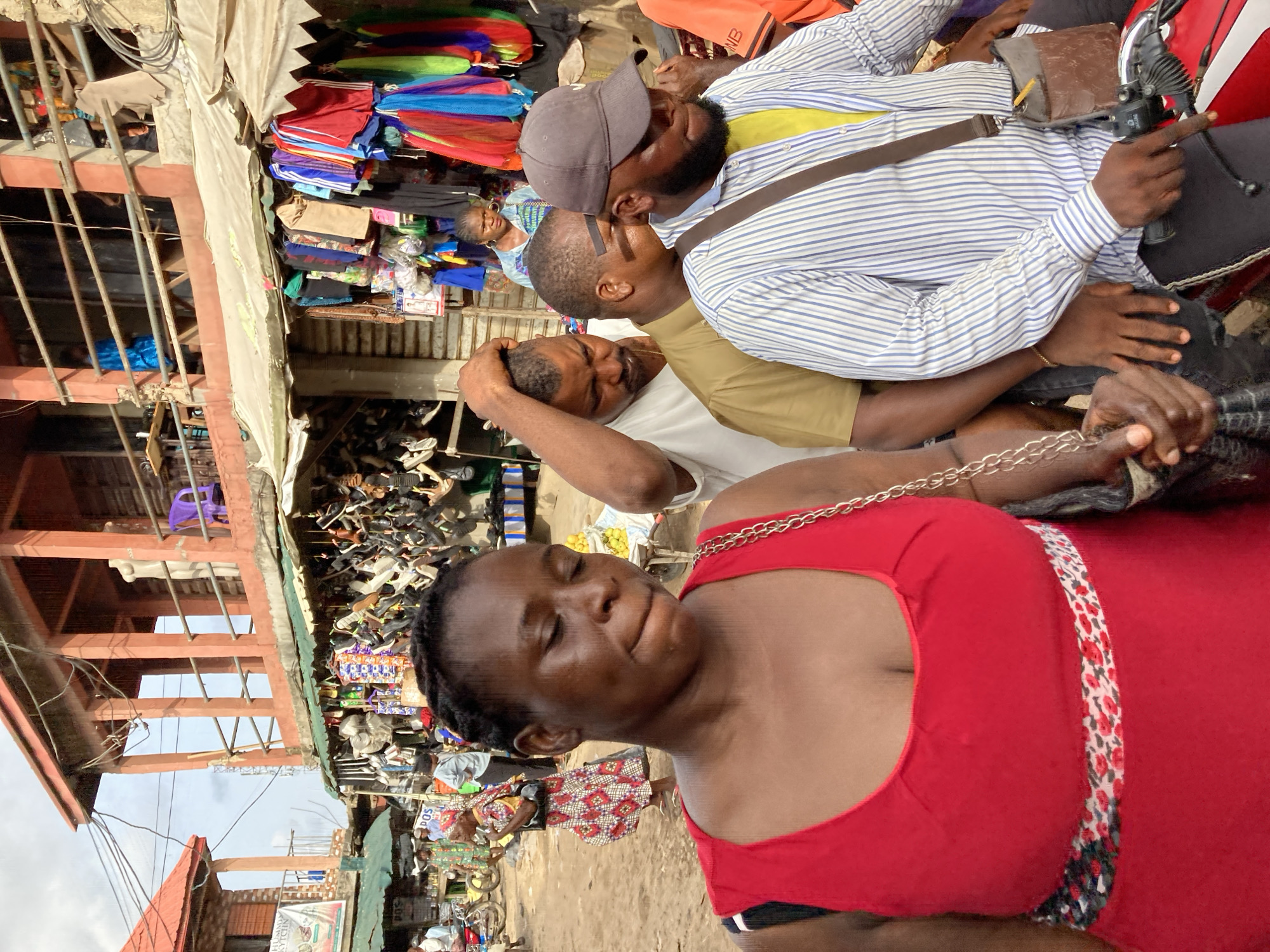 The image depicts a crowded marketplace in Ura Nta Umuarandu, Nigeria. A woman in a red top with a floral-patterned trim is prominently featured in the foreground. In the midground, three men are visible: one wearing a grey cap and a striped shirt, another in a tan shirt, and a third in a white shirt, all potentially riding a motorcycle. The background showcases a bustling marketplace with stalls displaying various goods, including shoes and clothing. Buildings with red roofs and metal structures are also visible. Overhead electrical wires run across the scene. The overall environment appears to be busy and congested, typical of a marketplace.