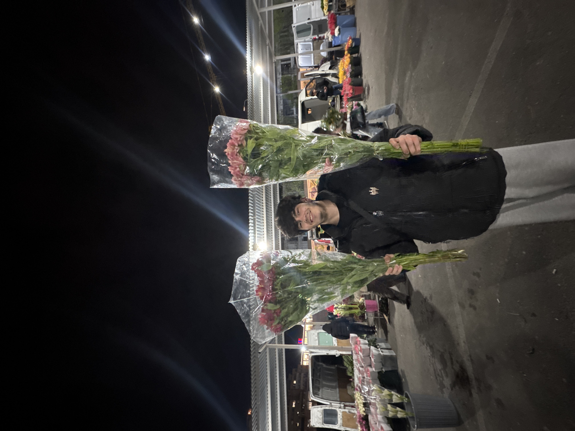A person stands at a flower market holding two bouquets of flowers. The person is wearing a black jacket and grey pants. The background shows a flower market at night. Several white vans can be seen with flowers being sold in front of them. Based on the streetlights and building design, this location is likely in Yerevan, Armenia.