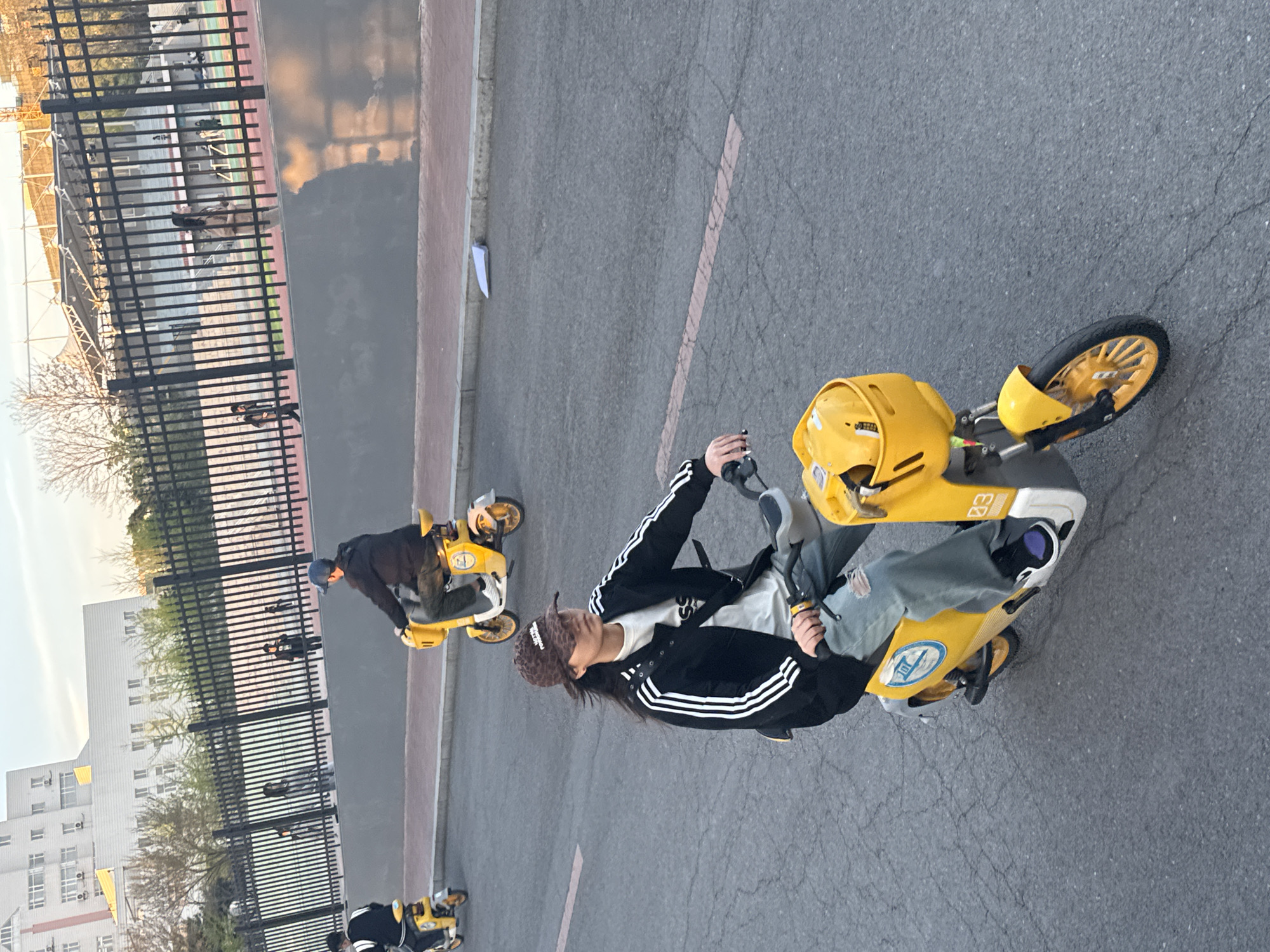 The image shows a street scene in Zhongtun, China, with several individuals riding yellow scooters. A person wearing a black Adidas jacket and a patterned hat is prominently featured on a scooter in the center-right of the frame. They are holding the scooter's handlebars. Additional people are riding similar scooters in the background. A fence runs along the left side of the image, with buildings visible beyond it. The road surface appears cracked and worn.