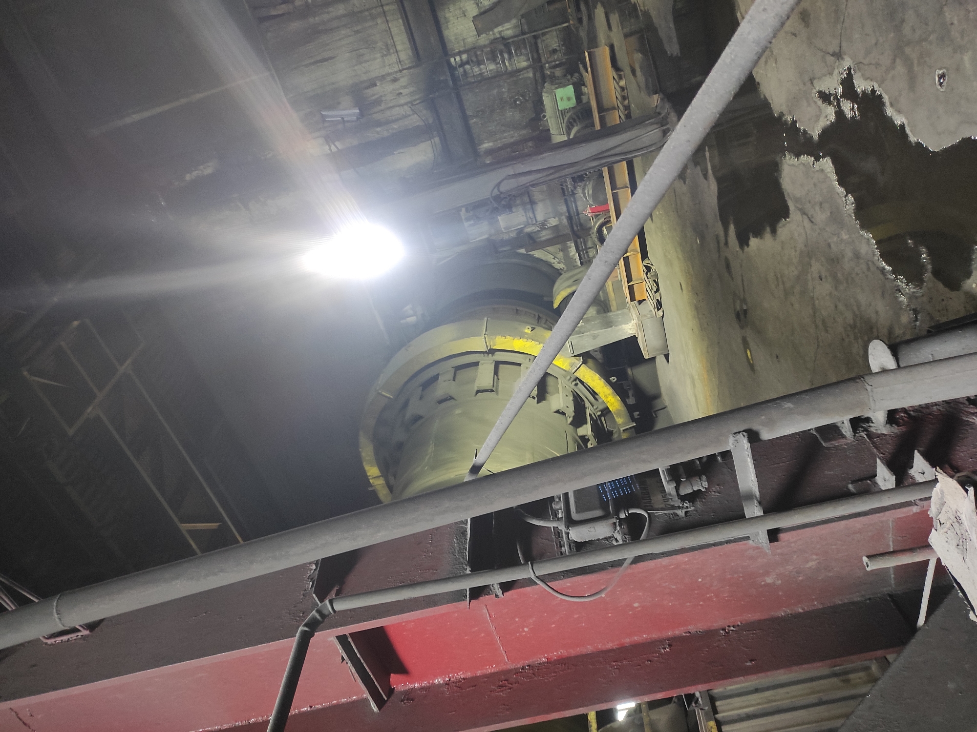 The image depicts the interior of an industrial facility, possibly a cement plant or similar manufacturing site, located in the city of Kyzyl, Kazakhstan. Dominating the scene is a large, cylindrical industrial machine, possibly a rotary kiln, with a yellow band around its circumference. In the foreground, a thick, red metal beam supports a grey pipe. The floor is concrete and wet in patches, indicating a humid or recently cleaned environment. Light sources, including a bright overhead light, cast strong reflections and lens flares across the image, obscuring some details. The overall atmosphere is dimly lit and industrial, with exposed pipes, electrical boxes, and various metal structures visible. There are no people present in the frame.