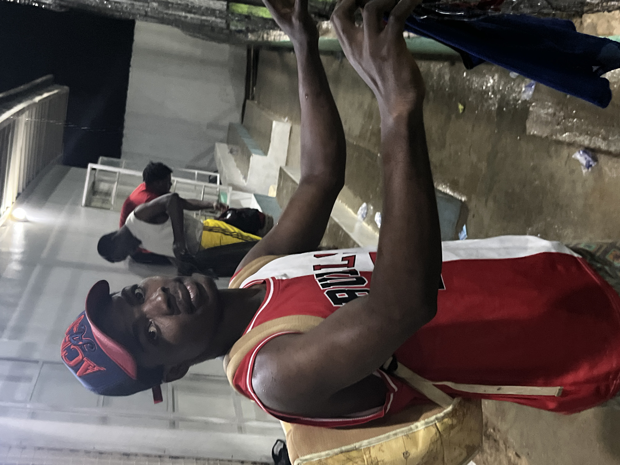 In an outdoor, dimly lit setting, possibly a sports facility or communal area, a young Black man is the primary subject. He is wearing a red and white sleeveless basketball jersey with "BULLS" printed across the front, a blue and red baseball cap, and a tan backpack slung over his left shoulder. His gaze is directed towards the camera, and he appears to be holding onto a dark blue garment with his right hand, which is extended towards the right edge of the frame. The ground is wet and appears to be concrete, with some debris scattered around. In the background, other individuals can be seen, one in a red shirt and another in a white tank top and yellow shorts, suggesting a shared activity or gathering. The environment features a covered overhang with lighting, and what appear to be tiered seating or structures in the background. The scene conveys a sense of activity and perhaps post-event or casual interaction. The location of Jos, Nigeria, is identified as the setting for this image.