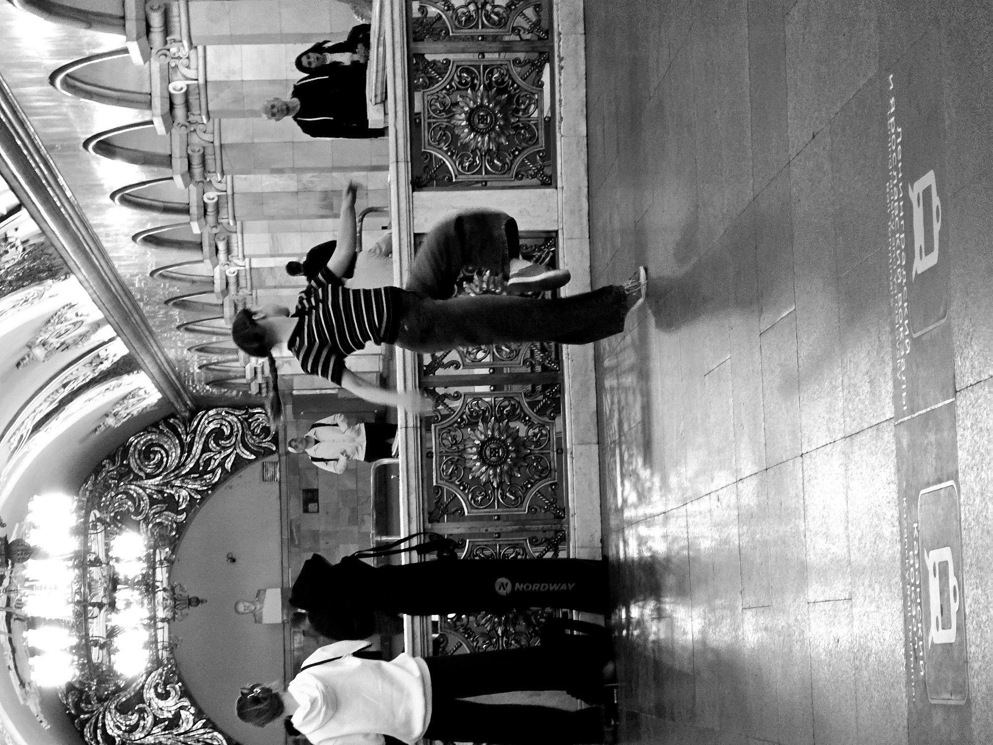 The image depicts the interior of a Moscow, Russia metro station. The scene includes several people. One person, dressed in a striped shirt and dark pants, is leaping mid-air. Other people are standing, walking, or otherwise occupying the space. One person is wearing a white shirt with suspenders.
The environment includes ornate archways, decorative metallic door panels, and tiled flooring with text and graphic symbols denoting the Kazan railway terminal. A Nordway branded bag is visible. The lighting appears to be a combination of natural and artificial, with a large chandelier providing illumination.