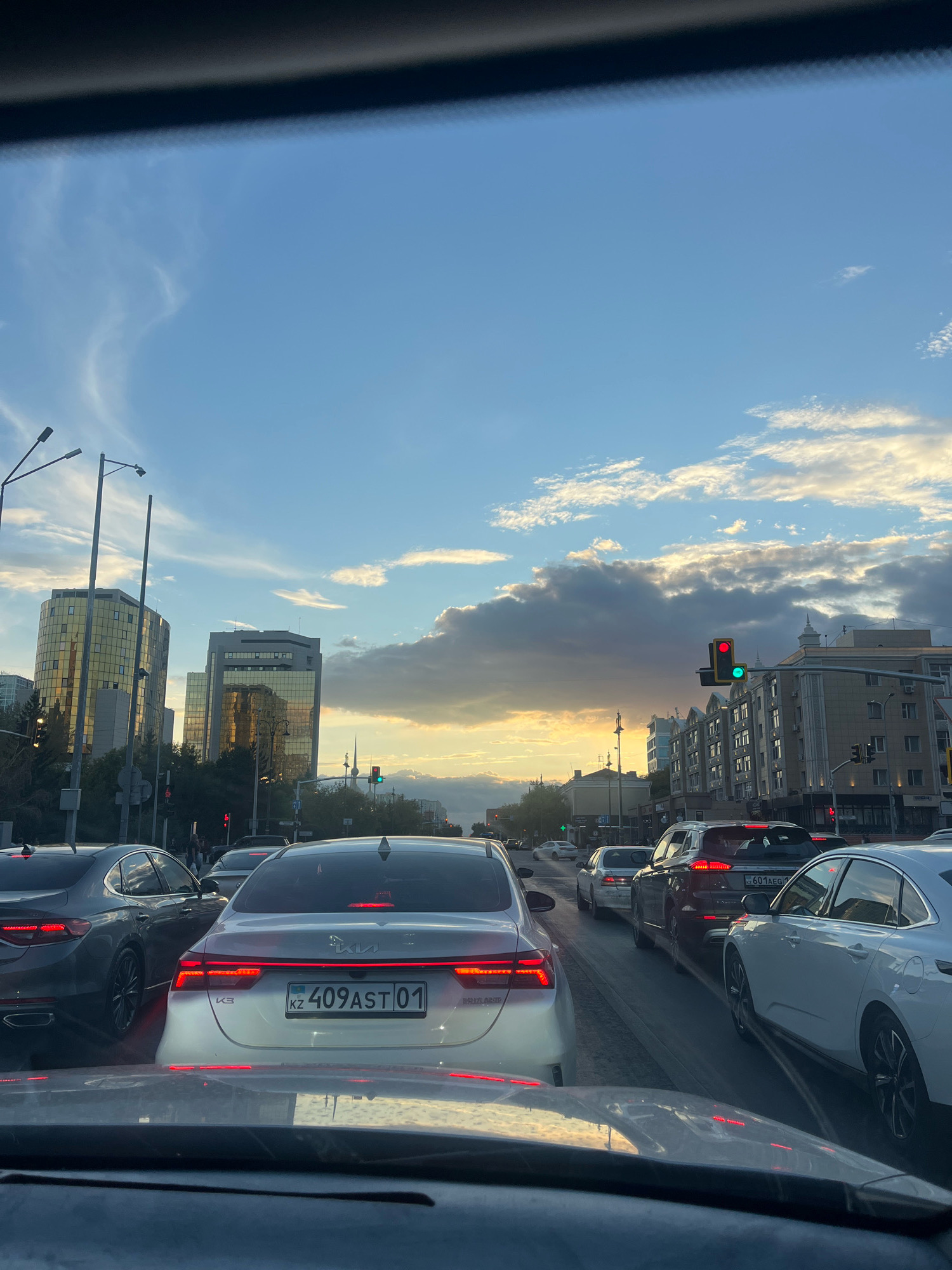 The image shows a view from inside a vehicle, looking forward at a city street during sunset. The sky is a mix of blue and orange with scattered clouds. In the distance, modern high-rise buildings with glass facades reflect the sunset light. Several lanes of traffic are visible, with numerous cars stopped, indicating a traffic jam. Traffic lights are present, with one showing red and green. Streetlights and poles are also visible along the sides of the road. The overall scene depicts a busy urban environment in Astana, Kazakhstan, during the evening commute.