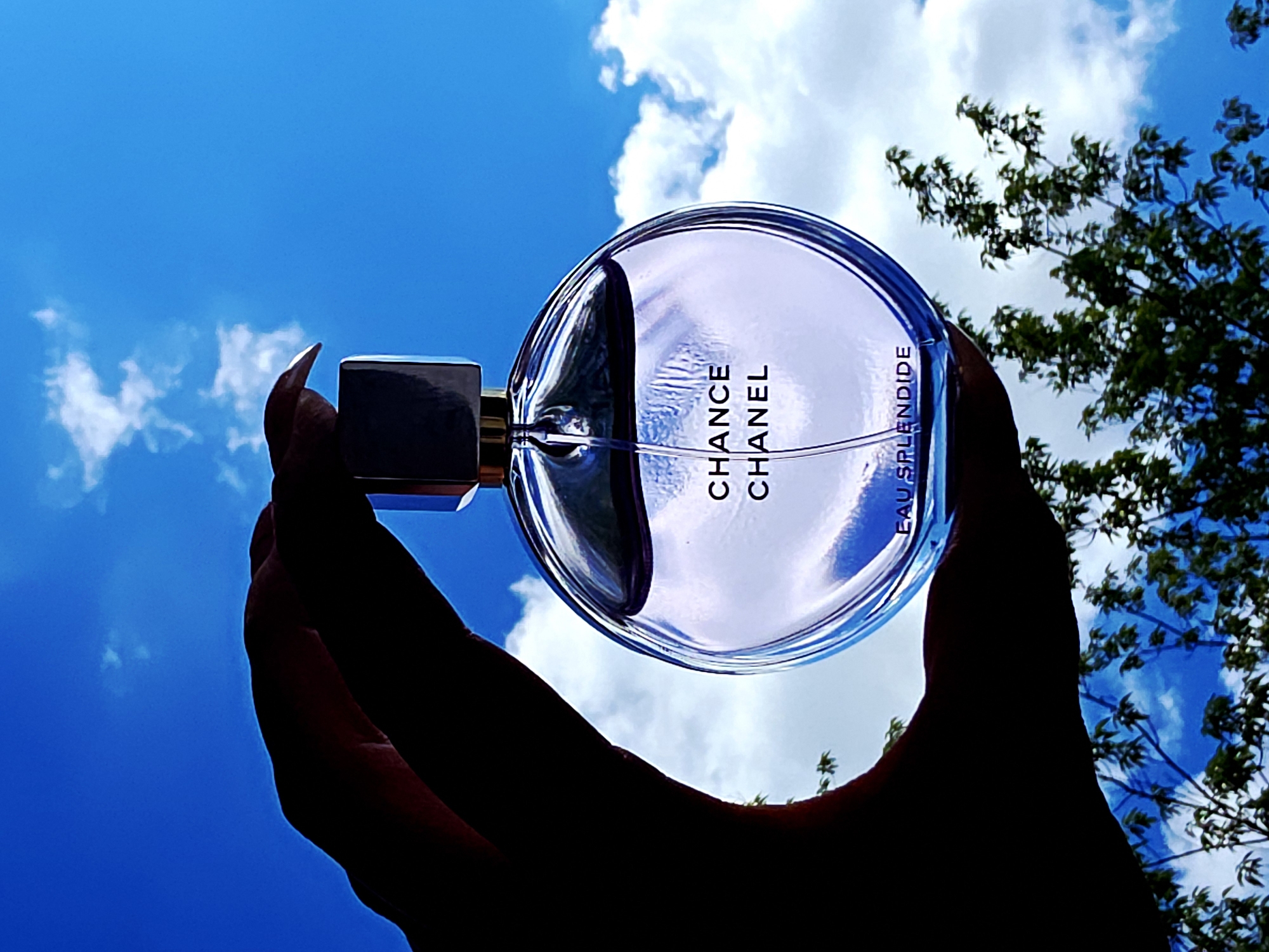 A low-angle shot captures a silhouetted hand holding a round glass perfume bottle against a vibrant blue sky dotted with scattered white clouds. The hand and the bottle's cap appear dark, creating a strong contrast with the bright background. The glass of the bottle is translucent, reflecting the blue sky and white clouds, giving it a luminous quality. Vertically oriented text is visible on the bottle, reading "CHANCE CHANEL" in prominent letters, and below it, "EAU SPLENDIDE" in smaller print. To the right, dark green foliage from a tree is visible, framing the scene. The image depicts a clear, sunny day outdoors.