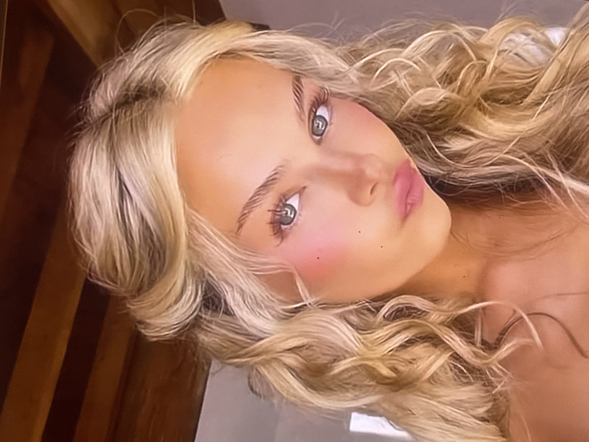 A close-up, angled portrait shows a young woman with light blonde, voluminous curly hair. Her head is tilted, and she looks directly into the camera with light-colored eyes, possibly green or hazel. Her eyelashes are long and well-defined, complemented by neatly shaped eyebrows. She wears makeup, including pink blush on her cheeks and soft pink lipstick.
Her face takes up a significant portion of the frame, with her blonde hair framing her features and cascading around her head. The background is softly blurred and indistinct, featuring dark brown tones on the left, possibly indicating wood, and lighter, neutral shades on the right, suggesting an indoor setting. The lighting is soft and even, highlighting her facial features. The woman's expression is neutral and engaging. There is no visible text in the image. The overall scene suggests a posed photograph.