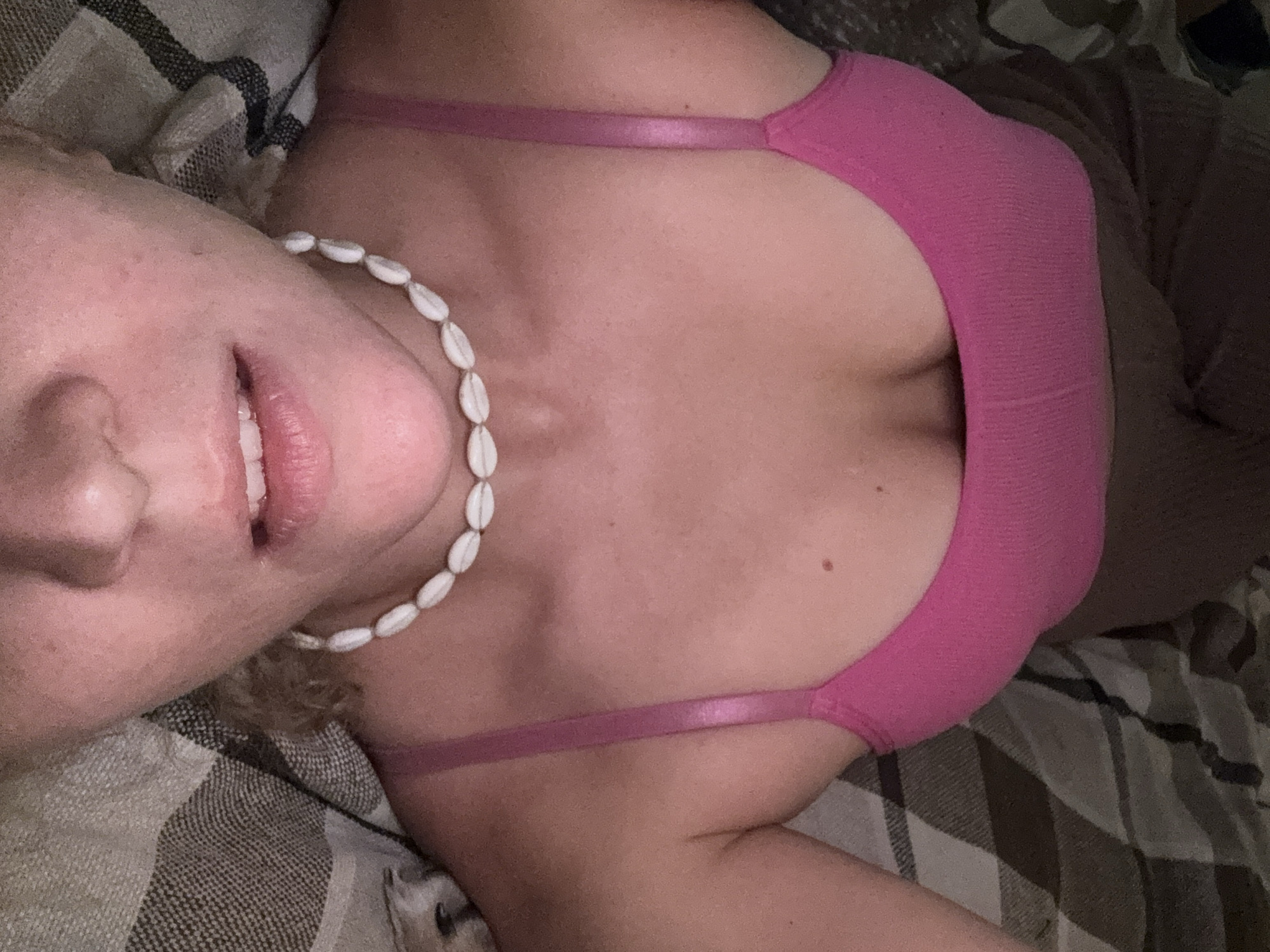 A woman is lying on a patterned fabric, possibly a blanket or bedding. She is wearing a pink top and a necklace made of white shells. Her face, including her nose and lips, is visible. The scene appears to be indoors. The location is not discernible from the image.