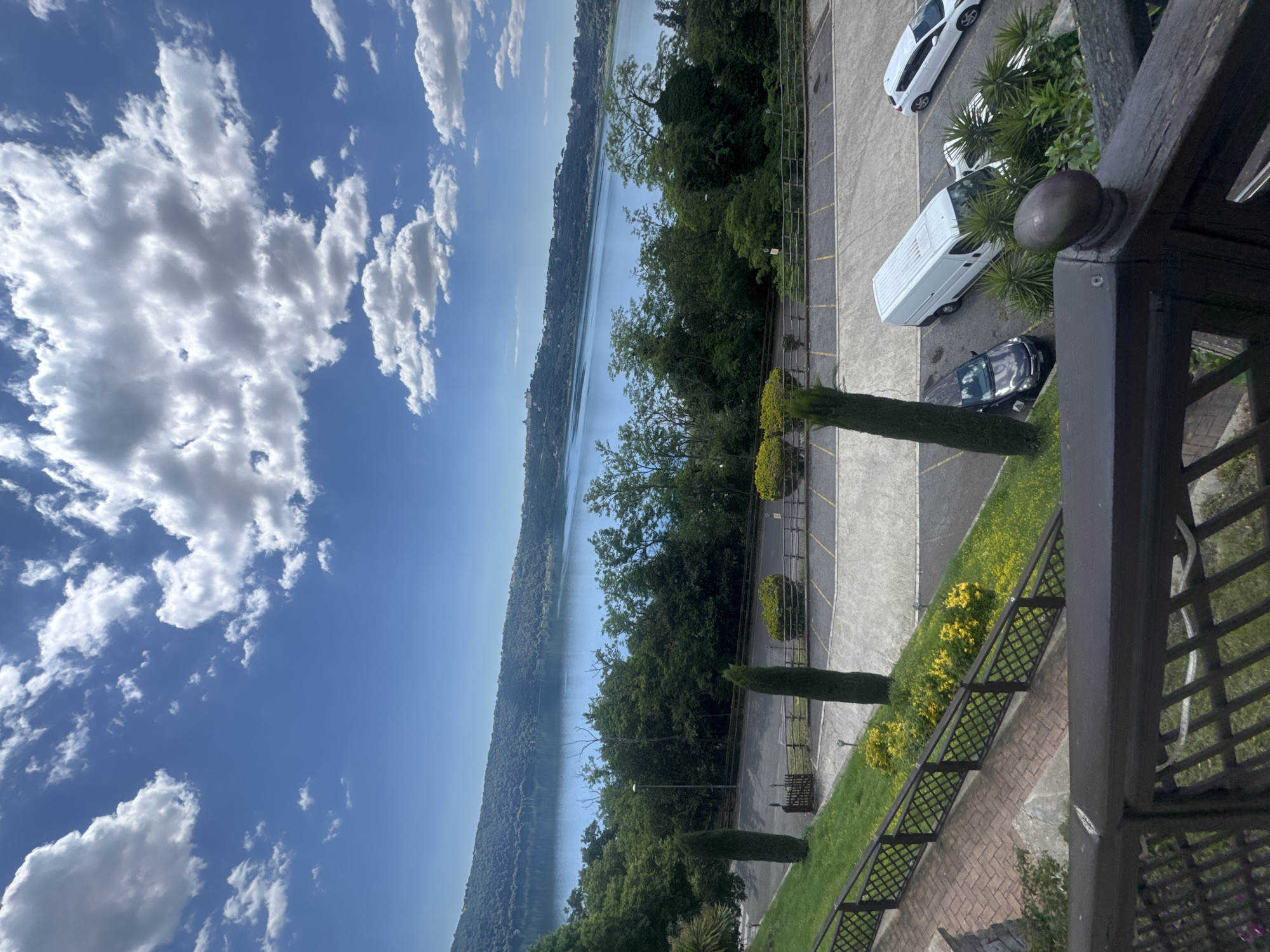 The image depicts a view overlooking Lake Albano in Castel Gandolfo, Italy. The sky is partly cloudy with a clear blue expanse. The lake fills a significant portion of the frame, bordered by tree-covered hills. Adjacent to the lake, there is a parking lot with several parked cars and a van. Manicured greenery and small trees line the edge of the parking area. A section of the balustrade from a balcony or terrace is visible in the foreground, suggesting the photo was taken from an elevated position.