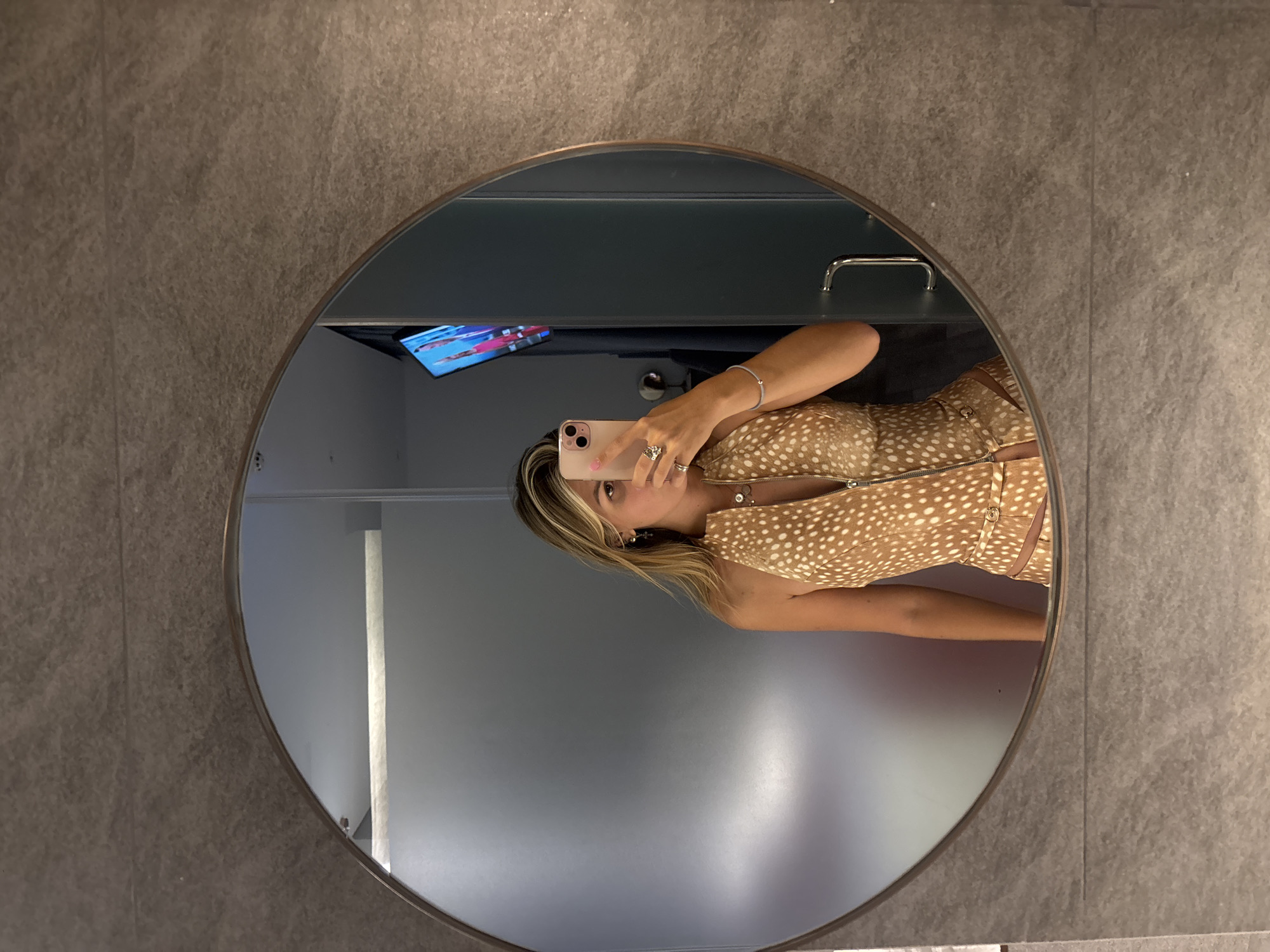 A woman with blonde hair is taking a selfie in a circular mirror. She is wearing a halter-neck top with a fawn print and a zipper down the front. She is also wearing rings and a bracelet. In the mirror's reflection, a television screen is visible on the wall behind her, displaying images of people. The background appears to be a modern bathroom with textured grey walls and a chrome handle on a door. The overall scene is indoors, likely in a hotel room or similar accommodation.
