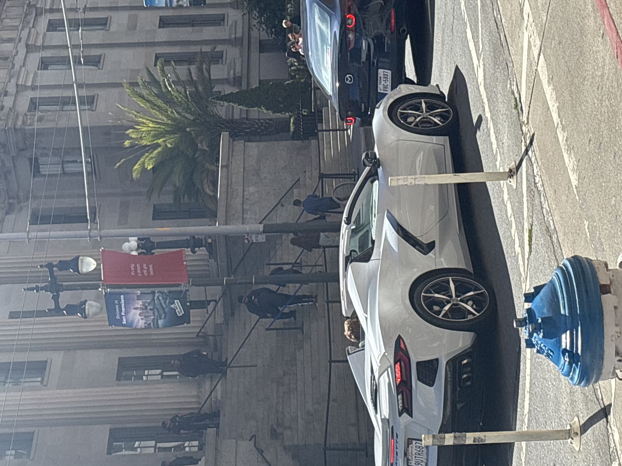 The image depicts a street scene in San Francisco, California, USA, with a white convertible sports car in the foreground, and a dark sedan behind it. A large, classical-style building with columns and tall windows serves as the backdrop. Several people are visible on the steps leading up to the building, some appearing to be in uniform. Banners hang from a lamppost in front of the building, one of which reads "Believe in San Francisco." A palm tree is also visible to the right of the building. The street has painted lane markings, and a blue fire hydrant is present in the lower part of the frame. The car in the foreground has its top down, and a person's head is visible inside. The license plate of the white car is partially obscured, showing "9UTR687". The license plate of the dark car reads "VHC-5807". The sun is shining, casting shadows on the street and pavement.