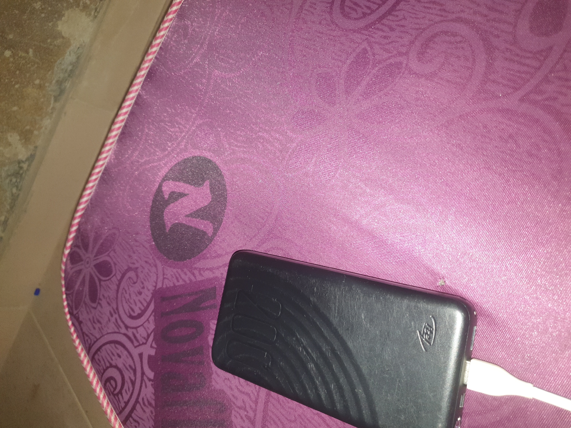 A black portable charger is connected to a white charging cable and lies on a pink mat with a patterned design. The mat has a pink and white striped trim. The charger has the embossed numbers "1000" and a small logo that appears to be "itel". The background is a textured, light-colored surface.