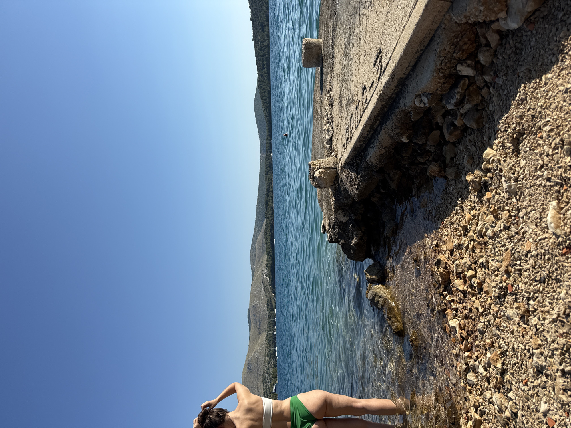 A person stands in shallow water by the shore, facing away from the camera and towards the sea. The individual wears a white bikini top and green bikini bottoms. Their hair is pulled back. The shoreline is rocky, with clear water revealing the seabed. Beyond the shore, the calm sea stretches to a tree-covered hilly coastline under a clear blue sky. Several sailboats are visible in the distance on the water. To the right of the frame, a concrete pier juts out into the water, with two stone posts on its surface. The pier appears weathered and in disrepair. The scene is likely located in Grad Šibenik, Croatia, a coastal region known for its numerous islands and clear waters.