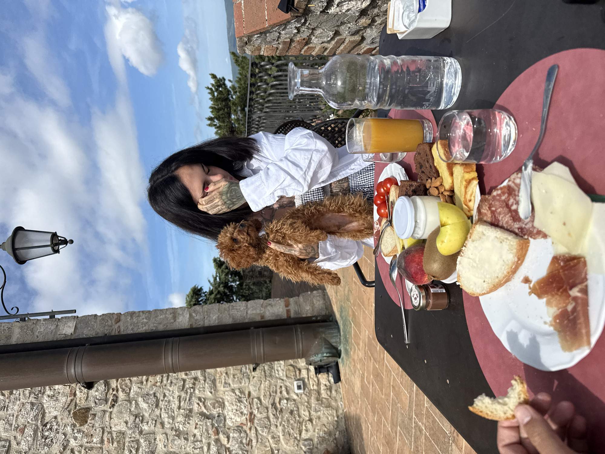 An outdoor breakfast or brunch scene unfolds on a terracotta-tiled patio in Castiglione d'Orcia, Italy, under a blue sky dotted with scattered white clouds. A woman with long dark hair, wearing a white oversized shirt over a gingham top, is seated at a dark table. She cradles a small, light brown, fluffy dog in her right arm. Her tattooed left hand, with red-painted nails, is raised to her face near her left eye.
The table is laden with a variety of food items, arranged on a red circular placemat. These include slices of bread (one buttered), an assortment of cured meats (salami, prosciutto) and cheese, small red tomatoes, a peach, a kiwi, a yellow pear, almonds, various pastries or cakes, a small jar of chocolate spread, and a white container of yogurt or cream. Beverages include a tall glass of orange juice, a clear glass bottle of water, and a smaller glass of water. A tanned hand in the lower left holds a bitten piece of bread, indicating the presence of a second person.
The background features a rustic stone wall on the left, against which a large, dark brown metal drainpipe with two banded sections runs vertically. Below it, an older, circular metal object and a white electrical box are visible. On the right, another stone wall supports an ornate black metal railing, overlooking a vista of green trees and distant rolling hills typical of the Val d'Orcia landscape. An antique-style black metal street lamp hangs from an unseen structure above.