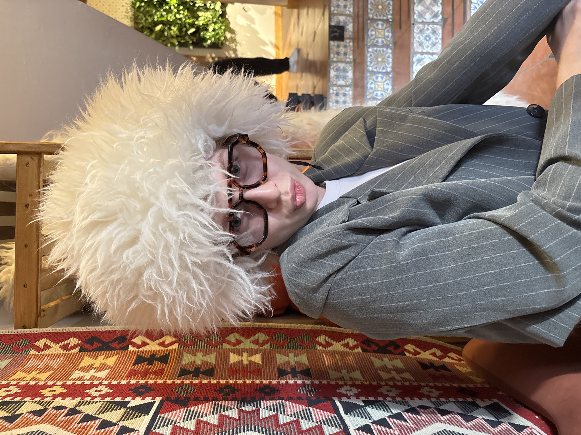 A person wearing large, tortoise-shell framed glasses and a large, fluffy white hat is lying on a patterned rug in an indoor space. They are also wearing a pinstripe grey suit jacket. The rug features geometric patterns in red, brown, yellow, and black. In the background are wooden furniture pieces and a wall with decorative tiles. The scene appears to be indoors, possibly in a home or a business with a unique design. There is no explicit information that this picture was taken in Perm, Russia.
